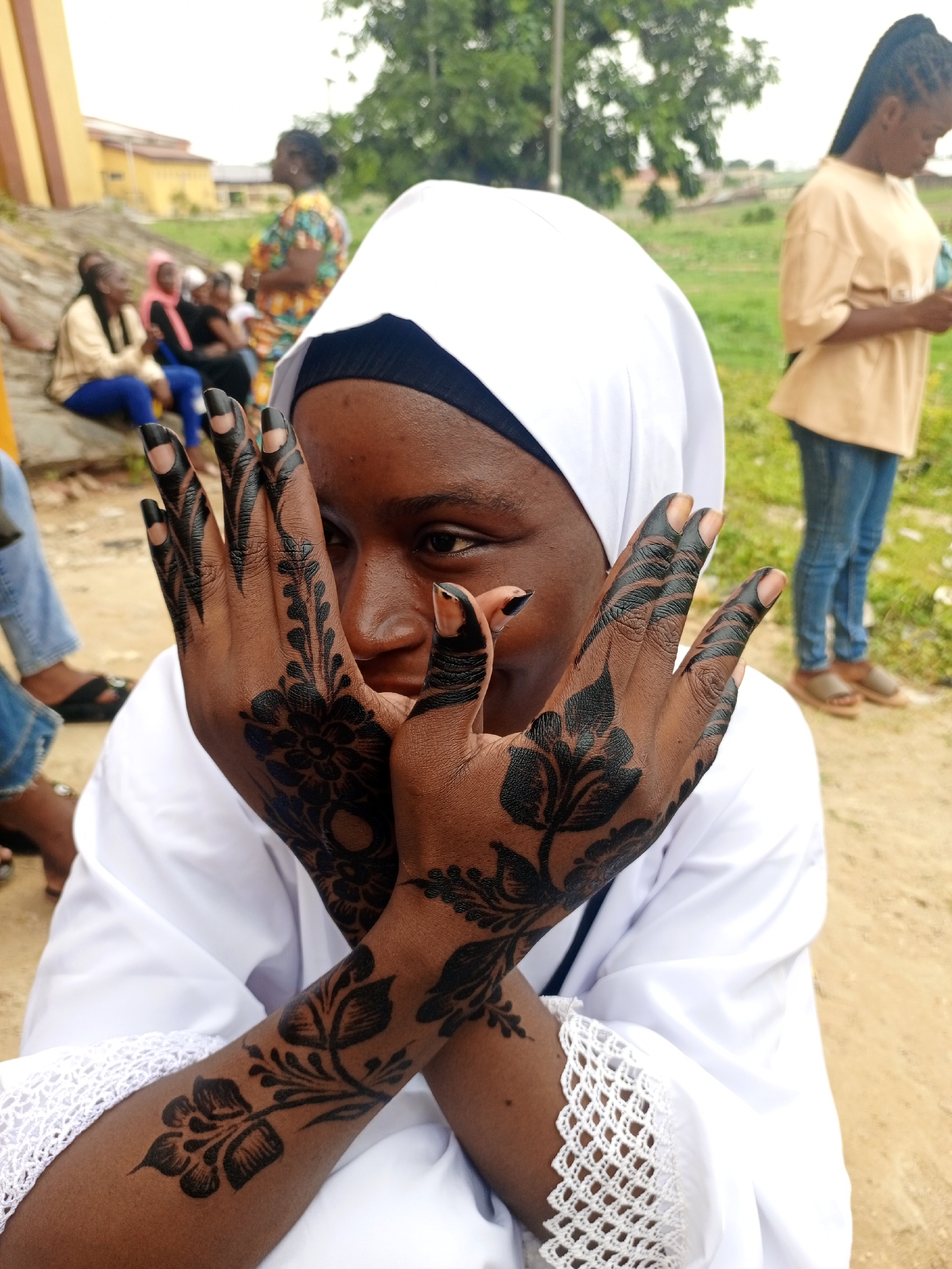 A person with brown skin and decorated hands covers their face with their hands. The person wears a white headscarf and white clothing with crochet detailing at the sleeve edge. Black henna-like designs cover the backs of the person's hands and fingers, extending onto the lower forearms. The person is centered in the frame.
In the background, other people are present. Some sit on a ledge to the left. A person in blue jeans and a yellow shirt stands to the right. A grassy area and some trees are visible behind them.
The location is likely in the city of Keffi, Nigeria.