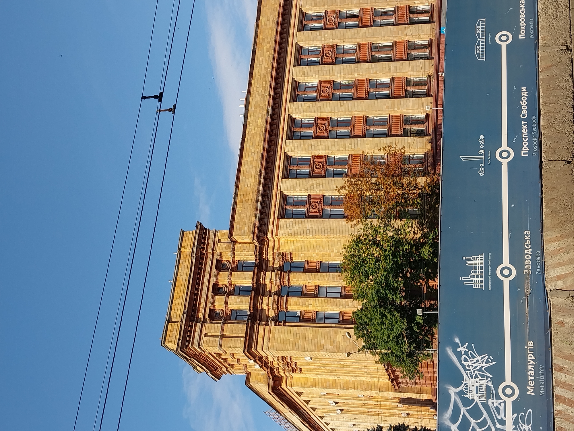 The image shows the facade of a large, multi-story building with a classical architectural style, constructed with light brown stone blocks. The building features numerous windows with dark frames, set within prominent vertical pilasters. The top of the building has a decorated cornice. In the foreground, a blue horizontal banner is visible, displaying graphics and text in white. This banner appears to be part of a public transportation route map or advertisement, with place names like "Металургія" (Metalurhiv), "Заводська" (Zavodska), "Проспект Свободи" (Prospekt Svobody), and "Покровська" (Pokrovska). The text indicates this is likely in the city of Dnipro, Ukraine, as suggested by the place names that correspond to metro stations in Dnipro. Several trees with green and autumnal foliage are situated in front of the building. Overhead, power lines stretch across the frame against a clear blue sky. The overall scene depicts an urban environment with historical architecture and public infrastructure.