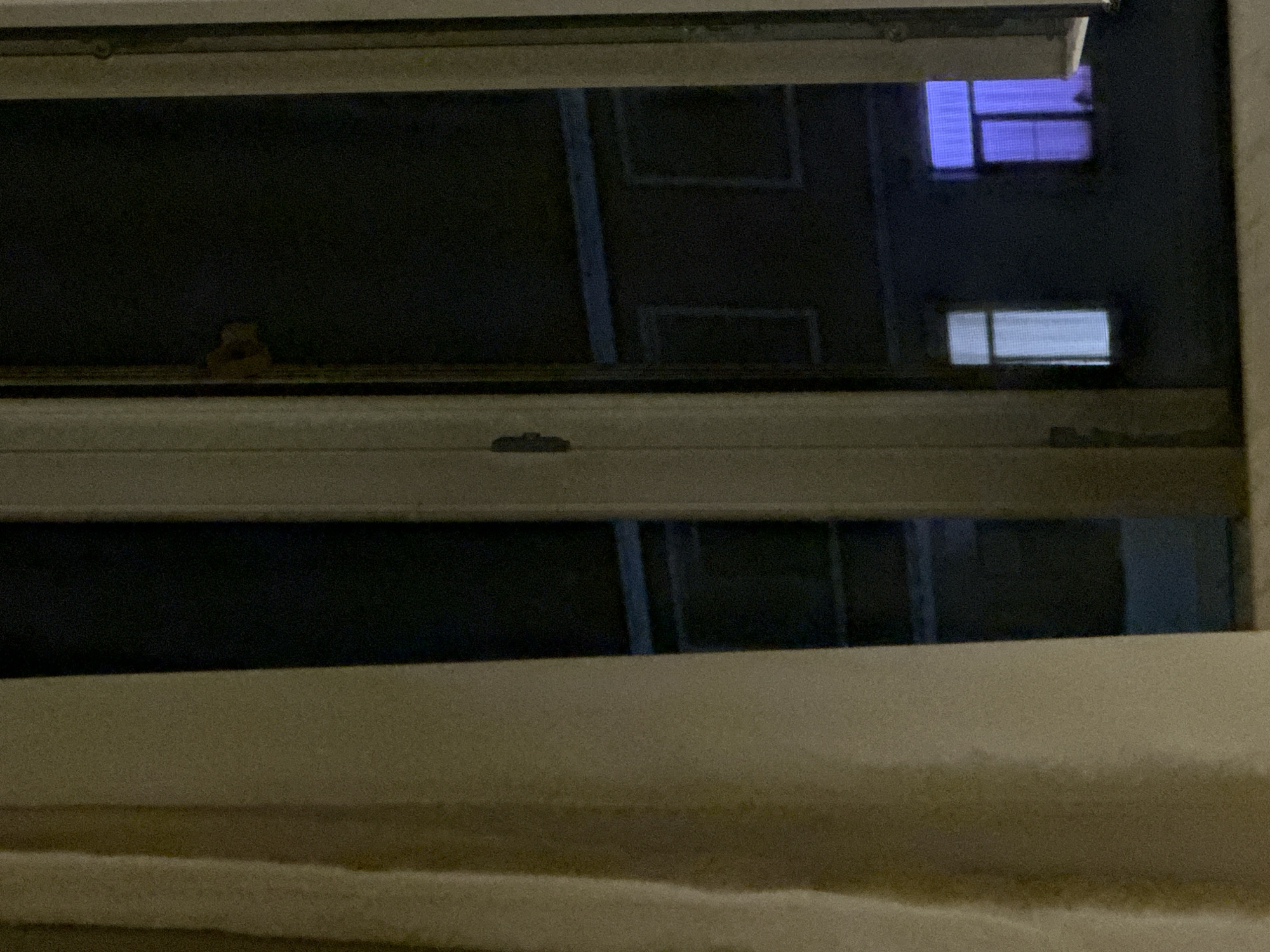 The image shows a view from a window at night, looking out onto a building with illuminated windows. A light-colored curtain is visible on the left side of the frame, partially obscuring the view. The window frame itself is visible, with vertical mullions dividing the panes. The exterior scene is dark, with reflections on the glass. Two windows on the opposite building are distinctly lit: one with a pale white light and another with a vibrant purple light. The overall impression is that of an urban residential setting at night. The image is undated and the specific location within Masazir, Azerbaijan is not determinable from the visual information alone. There are no people or discernible actions taking place.
