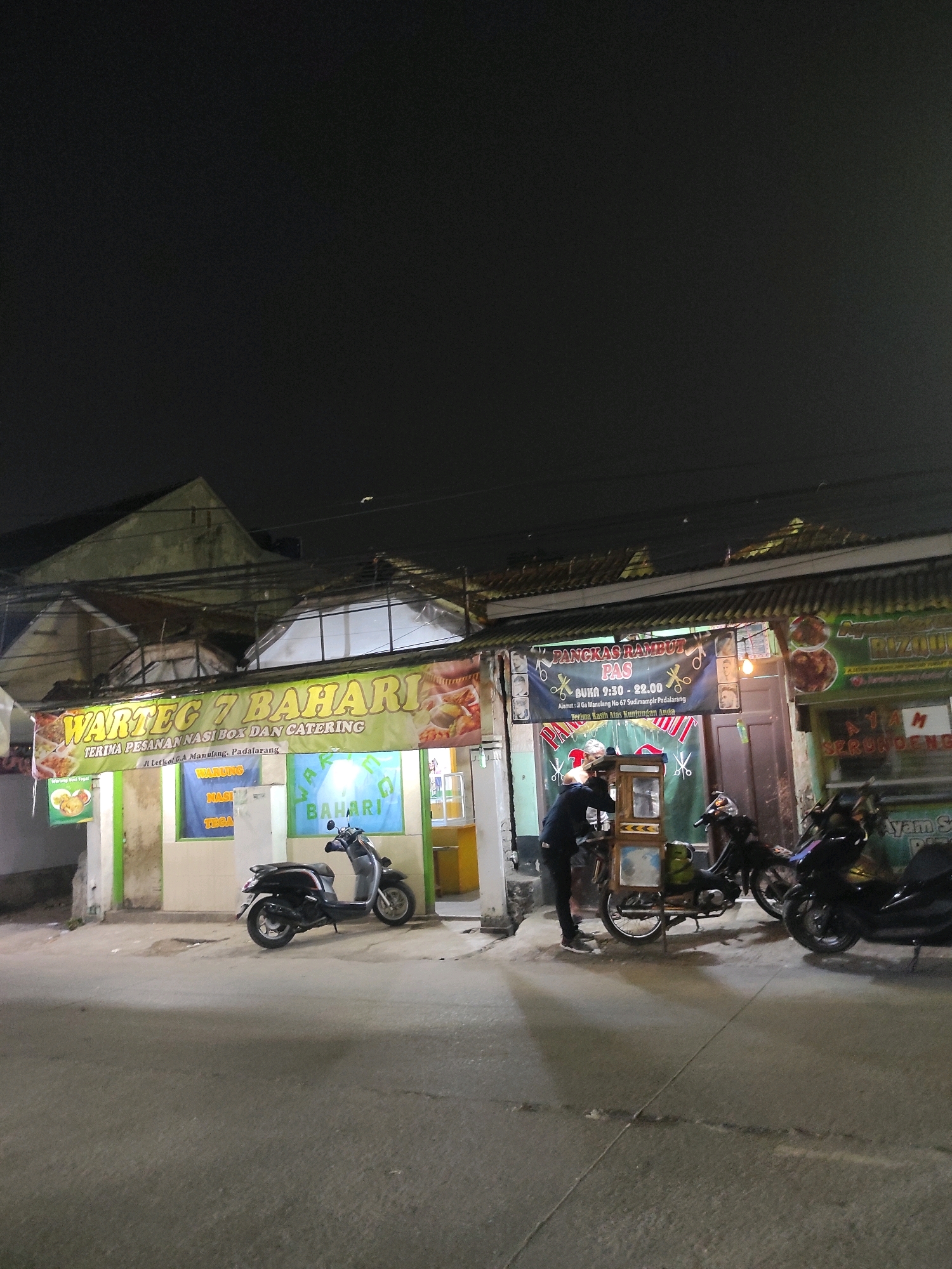 The image depicts a nighttime street scene in Padalarang, Indonesia. In the foreground, a paved street with visible expansion joints is present, casting long shadows. To the right, a dark-colored scooter is parked. Further to the right, a person in a dark jacket is interacting with a food cart. Behind them, another scooter, predominantly black, is parked. The background features several storefronts. A prominent banner on the left reads "WARTEG 7 BAHARI TERIMA PESANAN NASI BOX DAN CATERING" and provides an address: "Jl Letkol GA Manulang, Padalarang." To the right of this, another establishment has a banner advertising "PANGKAS RAMBUT PAS" (Haircuts PAS) with opening hours listed as "BUKA 9:30 - 22:00" and a similar address. The overall environment suggests a commercial area with food stalls and small businesses operating in the evening.