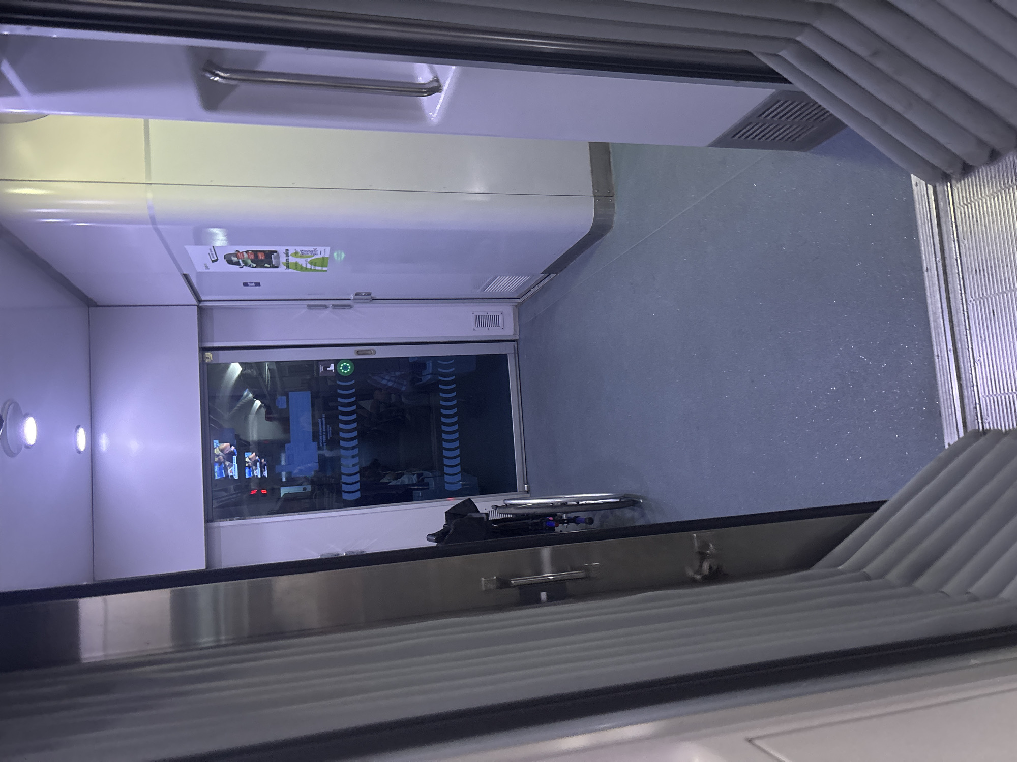The image depicts the interior of a train car, likely a passenger carriage, seen from an open doorway. The scene is brightly lit with overhead fluorescent lighting, casting a cool, bluish-white glow. The floor is a smooth, dark blue material with subtle sparkles.
In the foreground, to the left, is a partially open sliding door with a ribbed, white accordion-style curtain visible behind it. A metallic handle is attached to the door frame. To the right, another part of the train car interior is visible, including a section of a white wall with a vertical silver handle, and a silver grate vent near the floor.
The focal point of the image is the doorway leading further into the train car. Through the glass door, a hallway-like space is visible. On the left, a wheelchair is parked, its dark seat and chrome frame clearly discernible. The wheelchair appears to be empty. Beyond the wheelchair, through the glass, several passengers can be seen seated, their figures blurred by the distance and the glass.
On the right side of the hallway, a white wall continues, adorned with a colorful poster featuring an image of a car and text in a foreign language. A green circular indicator light is visible near the doorframe, possibly indicating an open or accessible status.
The environment suggests a modern public transportation setting. The overall impression is one of cleanliness and functionality. There is no information in the image to suggest a specific location in Bochmanivka, Ukraine, as it is a generic interior shot of a train car.