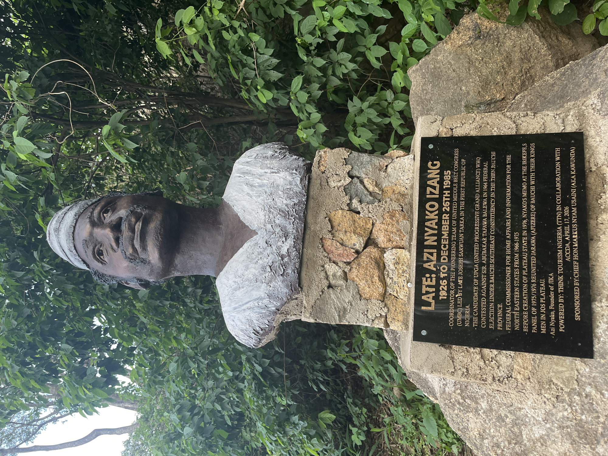 The image shows a bronze bust of a man, identified by an inscription as "LATE: AZI NYAKO IZANG 1926 TO DECEMBER 26TH 1985." The bust is mounted on a stone pedestal, which is situated on a larger rocky outcrop. The pedestal has a black plaque with white text detailing the achievements and affiliations of Azi Nyako. The background is filled with lush green foliage and trees, suggesting an outdoor, possibly natural or park-like setting. The inscription on the plaque mentions "JOS PLATEAU" and "JOS PLATEAU" again, indicating the location as being in Jos, Plateau State, Nigeria. The plaque also credits "THINK TOURISM NIGERIA (TTN)" for powering the initiative and "CHIEF HON. MARKUS NYAM USAINI (AKA KAWUNDA)" as the sponsor. The overall scene is a memorial dedicated to Azi Nyako.