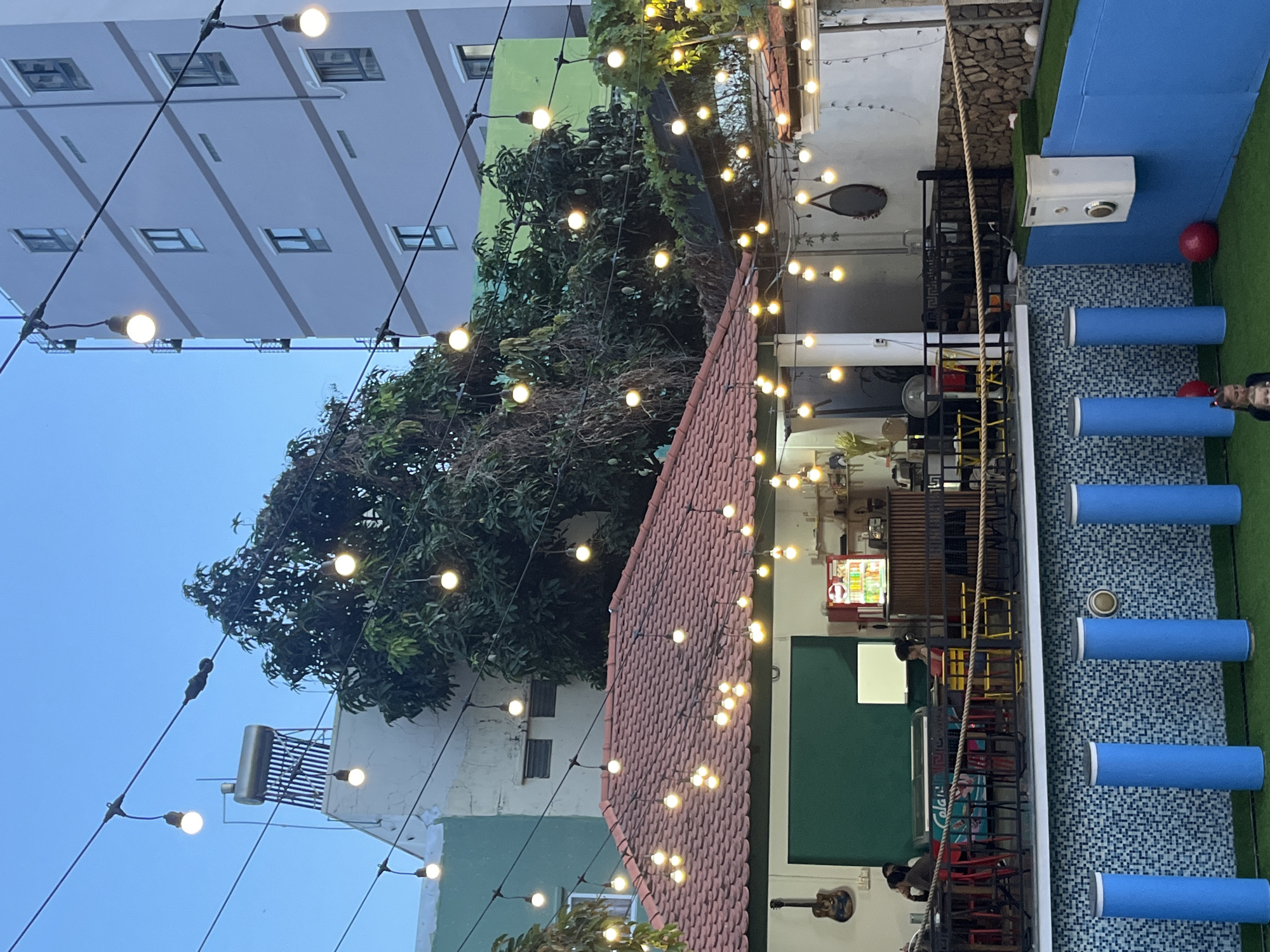 The image depicts an outdoor area in the city of Bac Nha Trang Ward, Vietnam. String lights are strung overhead. A building with a red tile roof and a green-painted exterior wall is visible, along with other buildings in the background. A tree is also present. There is an outdoor seating area with blue cylindrical cushions on a tiled surface next to a small area covered in green turf. A few people are visible in the lower right corner. There are tables and chairs near the red tile roof.