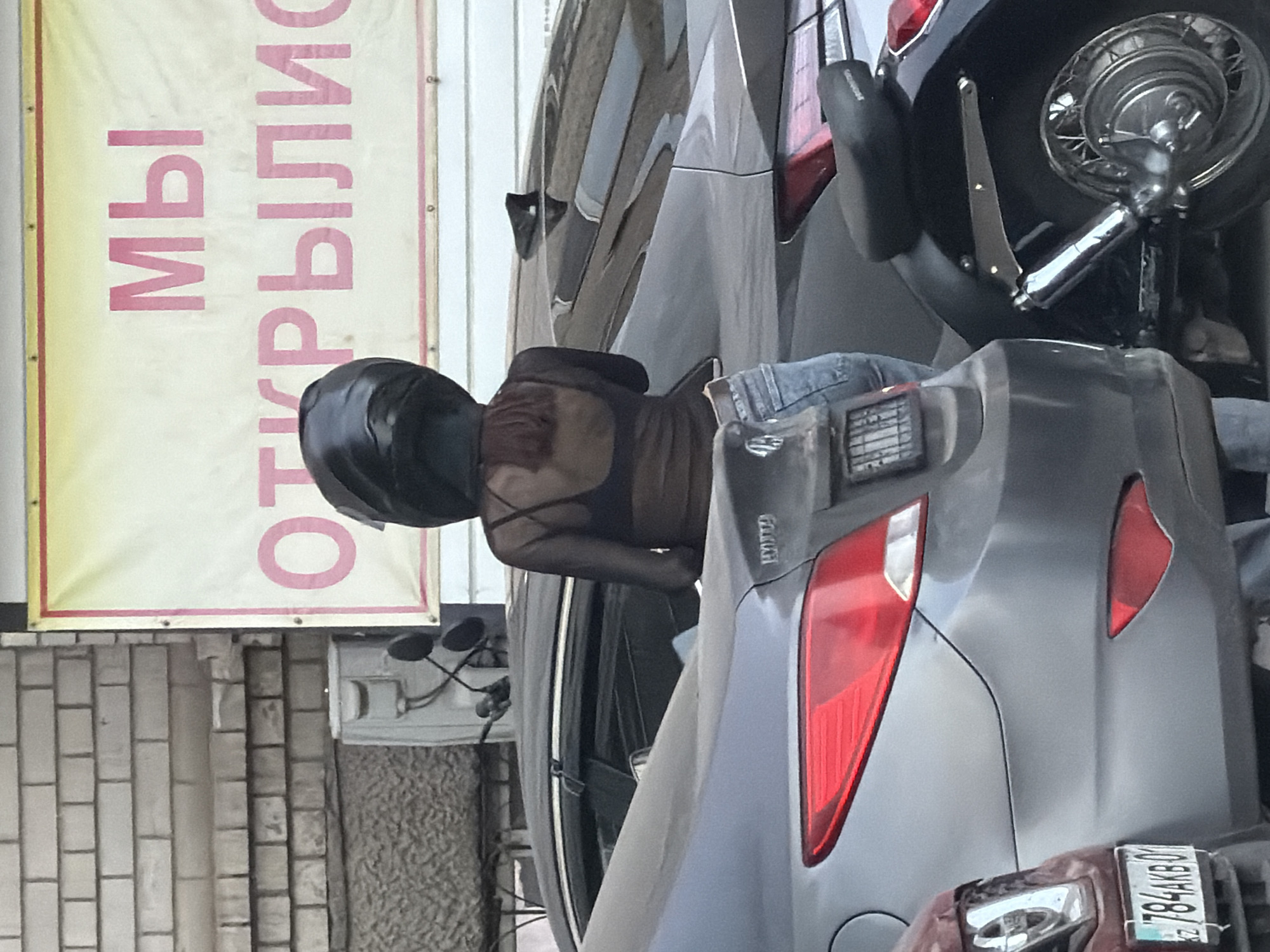 In Astana, Kazakhstan, a person wearing a black motorcycle helmet and a sheer black top over a black bra stands beside several parked cars and a motorcycle. The person is also wearing blue denim jeans. The scene appears to be in an outdoor setting, possibly a parking lot, with a brick wall and a large banner in the background. The banner has pink lettering in Cyrillic, which reads "МЫ ОТКРЫЛИСЬ" (We have opened). The license plates visible on the cars are in Cyrillic script, consistent with the format used in Kazakhstan. One visible license plate reads "784AKBO1". The lighting suggests it is daytime.