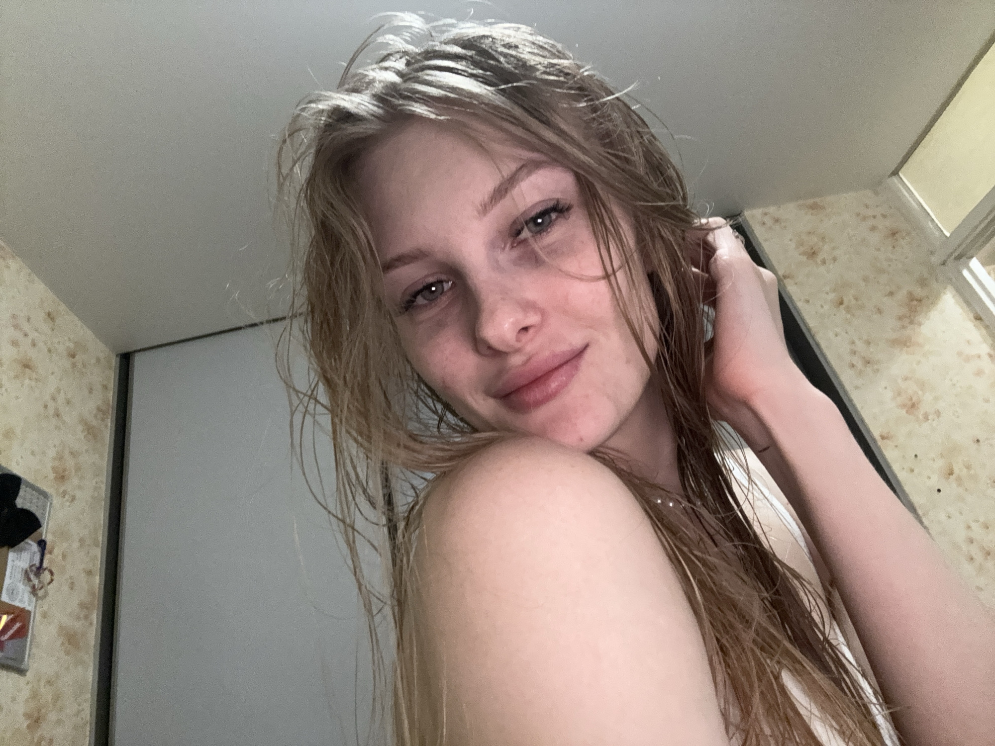 A young woman with damp, light brown hair and light-colored eyes poses for an indoor, close-up photograph, likely a selfie. She presents a soft smile, looking directly into the camera with her head slightly tilted. Freckles are visible across her nose and cheeks. Her left hand is raised to the left side of her head, appearing to touch or hold strands of her wet hair. She is wearing a light-colored, possibly white, top with a strap visible on her right shoulder. The background features a plain, light-colored ceiling. On the left, a wall covered in light brown speckled wallpaper is visible, adjacent to a plain light-colored wall or closet door, with a dark vertical frame line in between. Small, indistinct items hang on the speckled wallpaper. On the upper right, a light-colored, speckled countertop or shelf is present, bordering what appears to be a window frame or reflective surface. The specific location in the city of Perm, Russia, cannot be determined from the provided image.