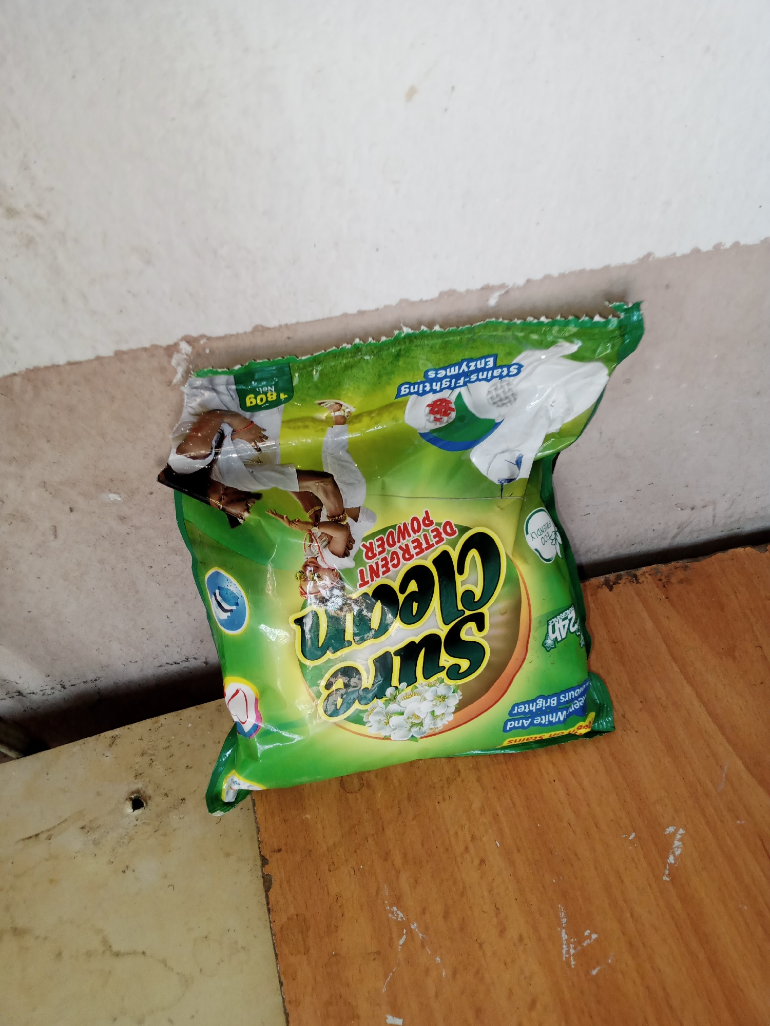 The image shows a crumpled green and yellow packet of "Sun Cleen Detergent Powder" on a wooden surface. The packet has white text and graphics, including flowers and a circular logo with a water droplet. The text "Stains-Fighting Enzymes" and "+80g Net" are visible on the top portion of the packet. The packet appears to be partially opened and is resting on a light-colored, textured surface that transitions to a wooden surface. The background consists of a textured, off-white wall with some grime and discoloration. The overall scene suggests an indoor setting, possibly a storage area or a cluttered surface. The image's composition and the presence of the detergent packet do not provide any direct indicators of the specific location within Jalingo, Nigeria, beyond being an interior space.