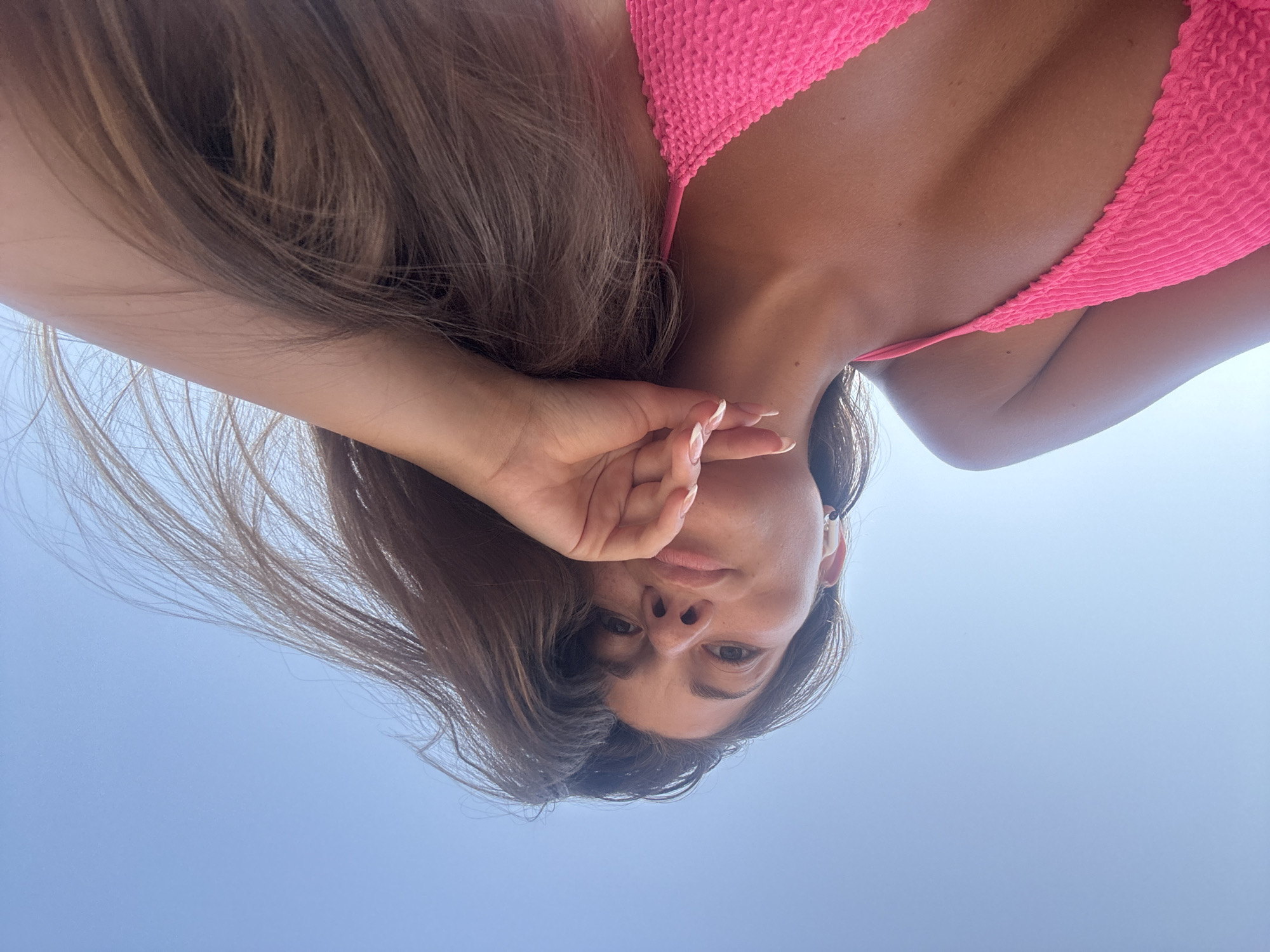 A woman wearing a pink bikini top is posing for a low-angle, selfie-style photograph against a bright, clear blue sky. She is looking directly at the camera with her head tilted and her hand partially covering her face. Her brown hair is partially obscuring her face. The location in Đà Nẵng, Vietnam is inferred due to the fact that it is an open blue sky that would be found at the beach.