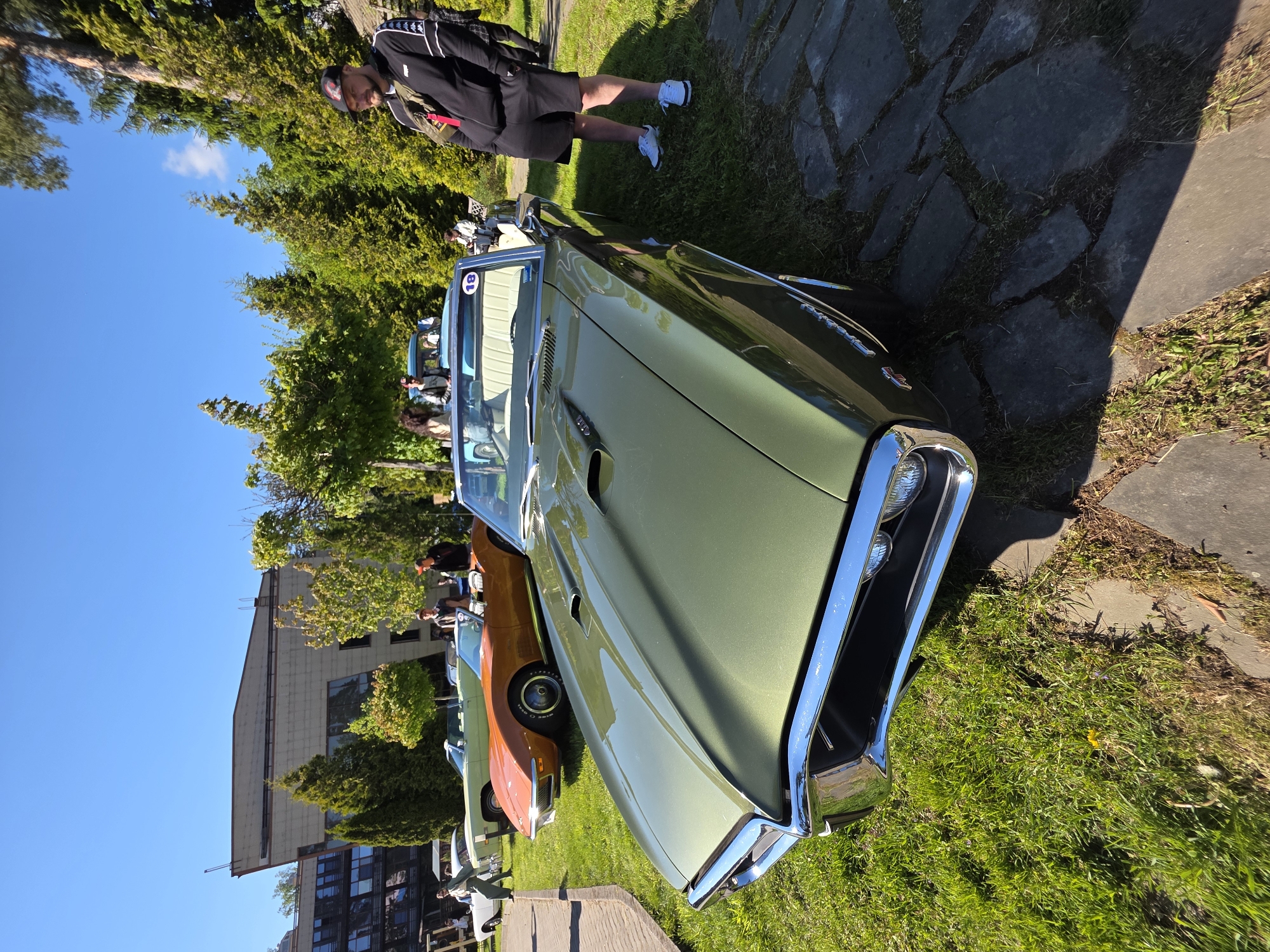 The image depicts a green classic convertible car parked on a grassy area, possibly in a car show or exhibition. A person stands nearby on a stone pathway, appearing to observe the vehicle. Other vintage cars are visible in the background, suggesting a gathering of classic car enthusiasts. A building is seen in the background. The location is Repino, Russia.