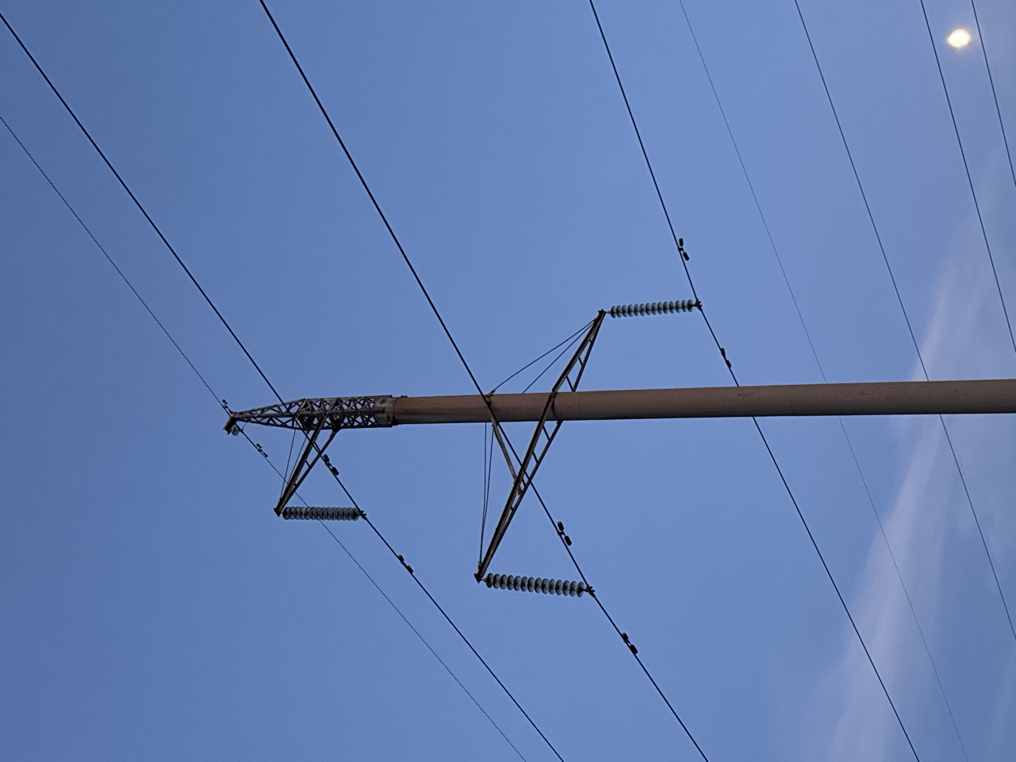 The image captures a low-angle shot of a tall, concrete power line pole against a clear blue sky. The pole is positioned in the center of the frame, extending upwards and partially out of view. Multiple electrical wires, supported by insulators, crisscross the sky. The insulators are visible as stacked, disc-like structures hanging from horizontal crossarms attached to the pole. The lower portion of the pole shows some rust-colored discoloration, suggesting age or exposure to elements. In the bottom right corner, a bright, circular object, possibly the moon or a distant light source, is visible. The overall scene suggests an outdoor, possibly rural or suburban, setting in Старое Михайловское, Russia. There are no people or animals present in the image, and no actions are depicted. The environment is characterized by the power line infrastructure and the expansive sky.