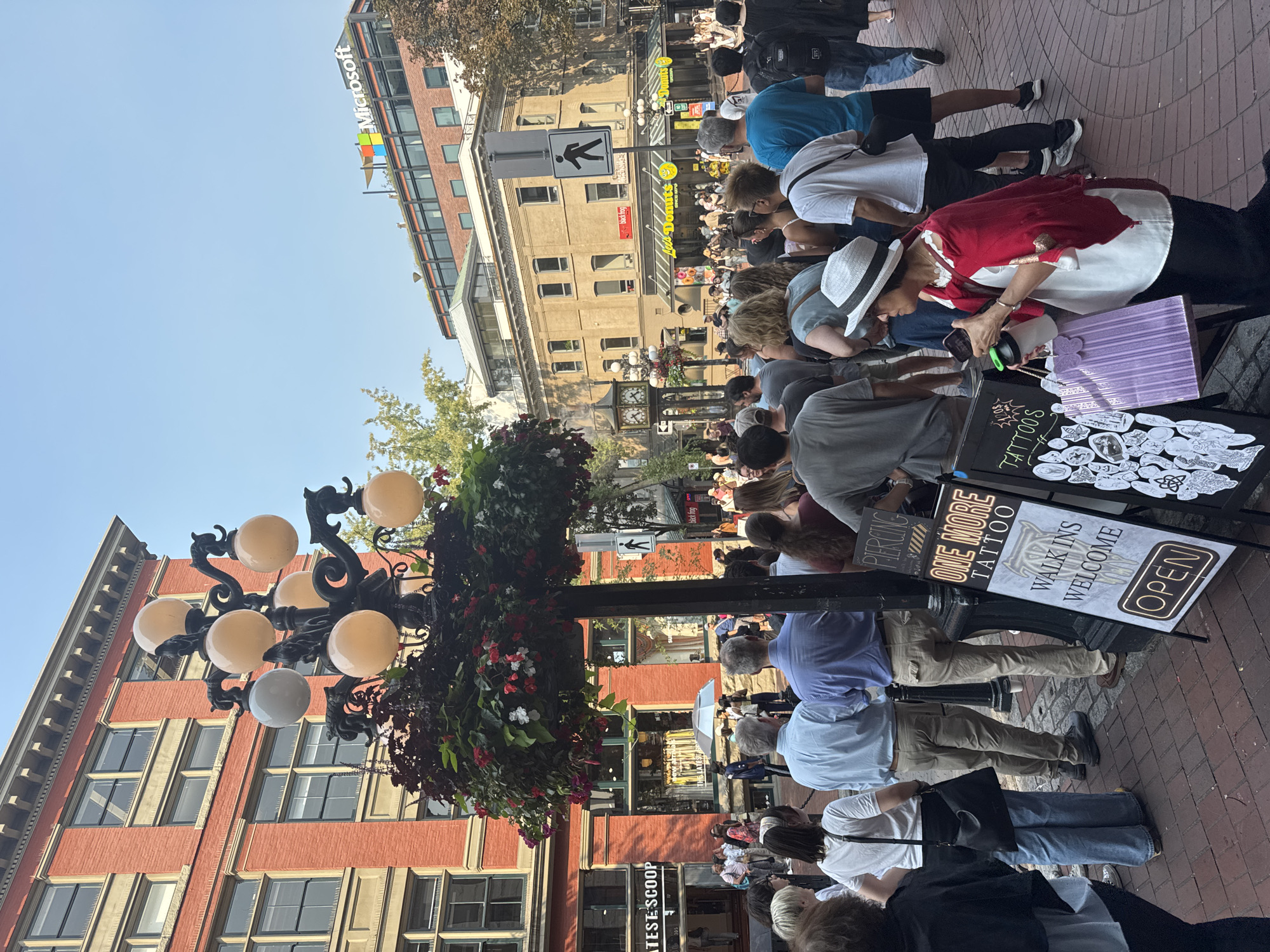 The image depicts a street scene in Vancouver, Canada, specifically Gastown, as indicated by the architecture and the presence of "The Latest Scoop" ice cream shop, a known establishment in the area. A crowd of people is gathered on a brick sidewalk. In the foreground, a decorative lamppost with hanging baskets of flowers stands prominently. To the right of the lamppost, two A-frame signs are visible, one advertising "ONE MORE TATTOO" with a "WALK INS WELCOME" message and an "OPEN" indicator, and another sign with various tattoo designs. People in the crowd are dressed casually, suggesting a warm day. Some individuals are looking towards the lamppost or the signs, while others are walking. In the background, buildings with red brick facades and modern architecture can be seen, including a building with a Microsoft logo. The overall atmosphere appears to be that of a busy urban environment on a bright, sunny day.