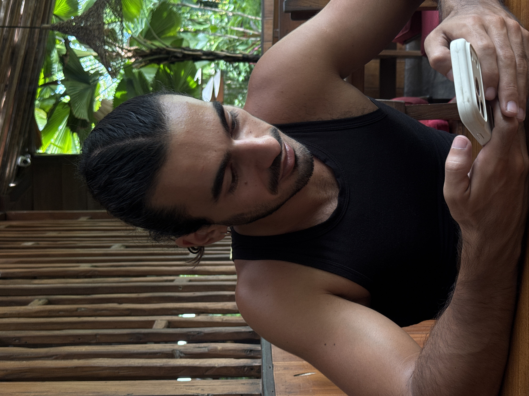 The image shows a young man horizontally across the frame, positioned in a relaxed manner. He has dark hair, styled in cornrows or braids, and a short beard. He wears a black tank top and has an earring in his left ear. His eyes are partially closed or cast downwards, giving him a calm, contemplative expression. In his hands, he holds a white smartphone encased in a white protective cover.
The man is leaning against or lying on a rustic wooden structure composed of parallel slats or logs, possibly a floor, wall, or elevated deck made of bamboo or unfinished wood. Light filters through the gaps in this structure, creating bright spots. In the background, beyond the wooden structure, there is an abundance of vibrant green tropical foliage with large, fan-shaped leaves, suggesting an outdoor or semi-outdoor setting in a lush environment. A blurry, light-colored netted or woven material is also visible in the upper background. The natural, soft lighting indicates it is daytime. The scene evokes a sense of relaxation in a tropical location, consistent with North Kuta, Indonesia. There is no visible text in the image.