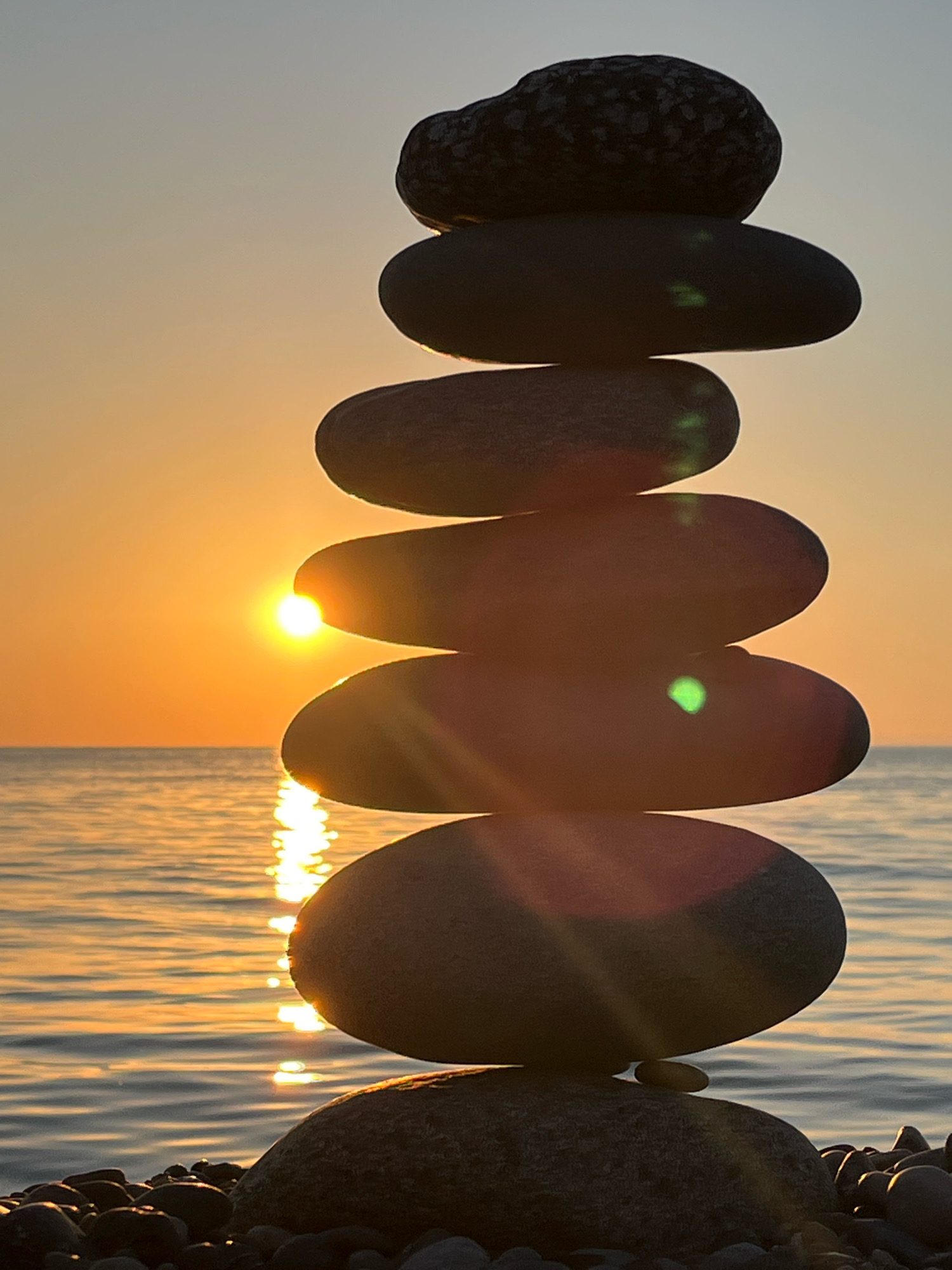 A stack of seven smooth, grey stones is balanced on a pebble beach at sunset. The sun, low on the horizon, casts a golden glow on the calm sea, creating a shimmering reflection. The stones are arranged in decreasing size from bottom to top, with the largest stone at the base and the smallest at the apex. The top stone is darker and more textured than the others. The sky is a soft gradient of orange and grey, suggesting the end of a day. The location is the beach in Kobuleti, Georgia. There are no people visible in the image.