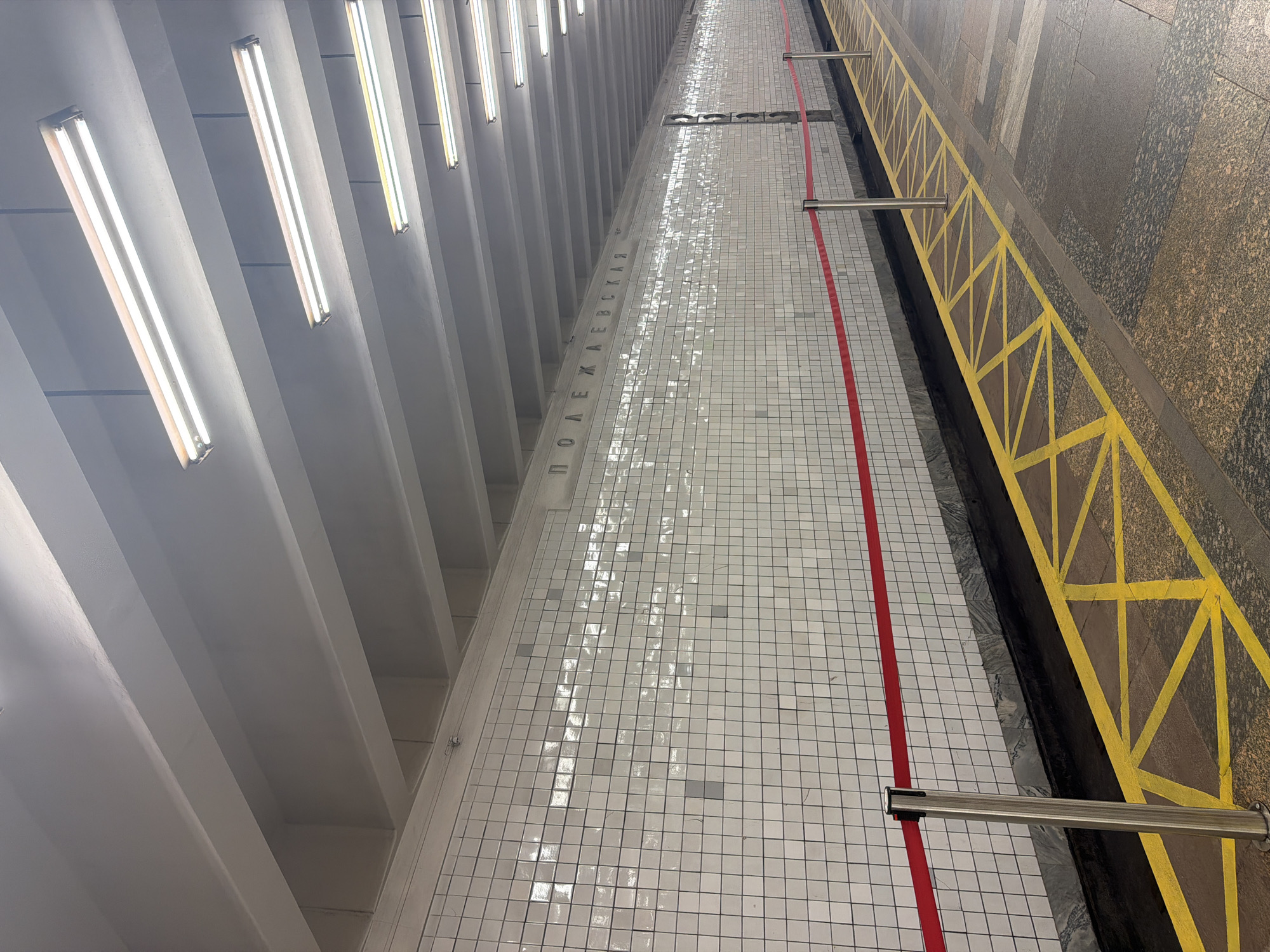 The image captures an interior view of a subway station, likely a platform. The scene is dominated by a long, tiled wall with the name "ПОЛЕЖАЕВСКАЯ" (Polezhaevskaya) spelled out in raised, metallic letters. The wall is primarily white tiled, with some tiles appearing darker or reflective, suggesting a patterned or textured surface, or possibly water damage. A bright red line runs along the platform edge, parallel to the wall, separated by a section of white tiled floor. To the right of the red line, a yellow safety barrier with a geometric pattern is visible, backed by a wall covered in dark, textured panels resembling granite. Fluorescent lights are mounted on the ceiling, casting a bright, even illumination throughout the space. The perspective is looking down the length of the platform, emphasizing its linear perspective and the repeating elements of the tiling, lights, and safety markings. There are no people visible in the frame. The overall impression is one of a functional, well-lit, and clean public space, typical of a metropolitan transit system.