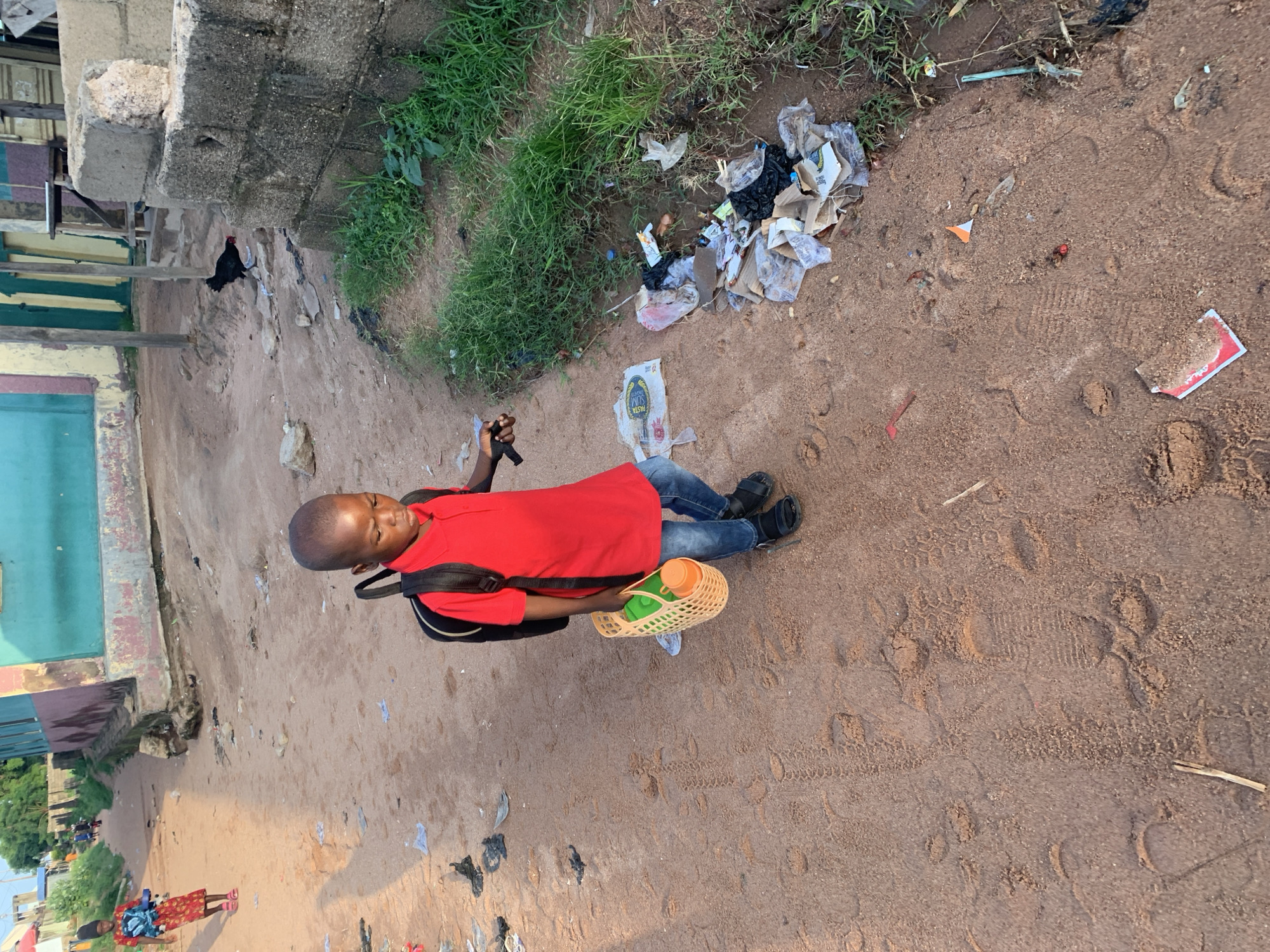 In Jalingo, Nigeria, a young boy in a red shirt and blue jeans walks along a dirt path. He carries a black backpack and a woven basket containing a green container and an orange bottle. The path is marked with footprints and scattered with debris, including plastic bags, paper scraps, and cardboard. To the right of the path, a pile of trash is visible near a patch of green grass. In the background, buildings with colorful facades line the street. A black chicken walks near a stone wall on the right side of the image. Further down the path, another person, possibly a child, is walking away from the camera, wearing a red and white patterned dress and pink sandals.