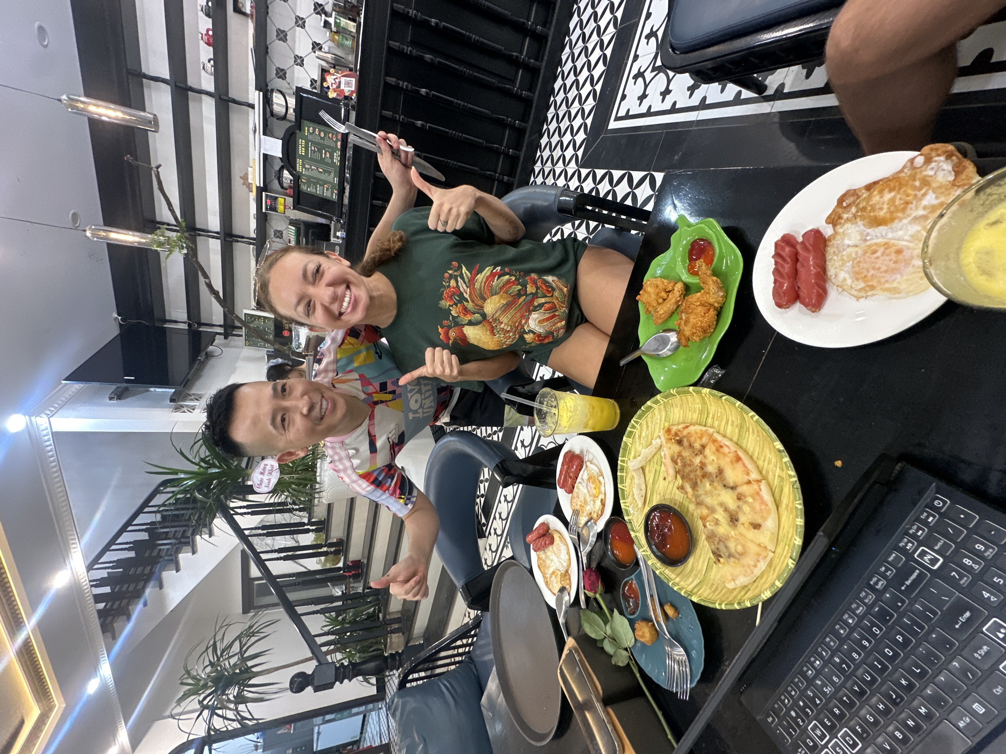 This image captures two individuals, a man and a woman, seated at a black table laden with food and drinks inside what appears to be a modern restaurant or cafe in Phố Ràng, Vietnam. Both subjects are smiling broadly and giving "thumbs up" gestures towards the camera, conveying a sense of happiness and enjoyment.
The woman, with light brown hair, is wearing a dark green t-shirt featuring a colorful rooster design and partially visible text "LOVE UNIT." The man, with short dark hair, is in a vibrant, multi-patterned shirt.
The table is filled with various dishes, including plates of fried sausages and fried eggs, a serving of fried chicken with ketchup, and a yellow, flatbread-like item (possibly a savory pancake or pizza) presented in a woven basket with a side of chili sauce. A glass of yellow juice is also visible, along with an open laptop, cutlery, and a single red rose, adding a touch of elegance or romance.
The background features a stylish interior with a striking black and white geometric tiled floor. A black wrought-iron staircase with potted plants ascends, suggesting a multi-level establishment. Modern pendant lights hang from the ceiling. A small sign near the man reads "Chúc Mừng Sinh Nhật," which is Vietnamese for "Happy Birthday," indicating a celebratory event. The laptop keyboard shows standard English QWERTY characters.