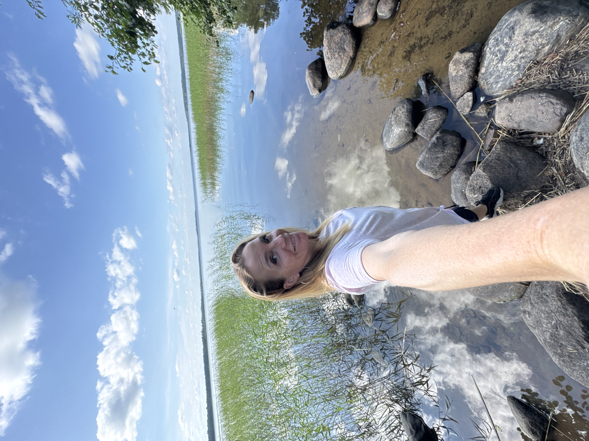 A blonde woman stands on the rocky shore of a lake, taking a selfie. She wears a pink t-shirt and black shorts. The lake is surrounded by tall green reeds, and the water reflects the blue sky and white clouds. A line of trees is visible in the distance. The woman is smiling at the camera. The overall scene is peaceful and natural. This image was taken in Yablonovka, Russia.