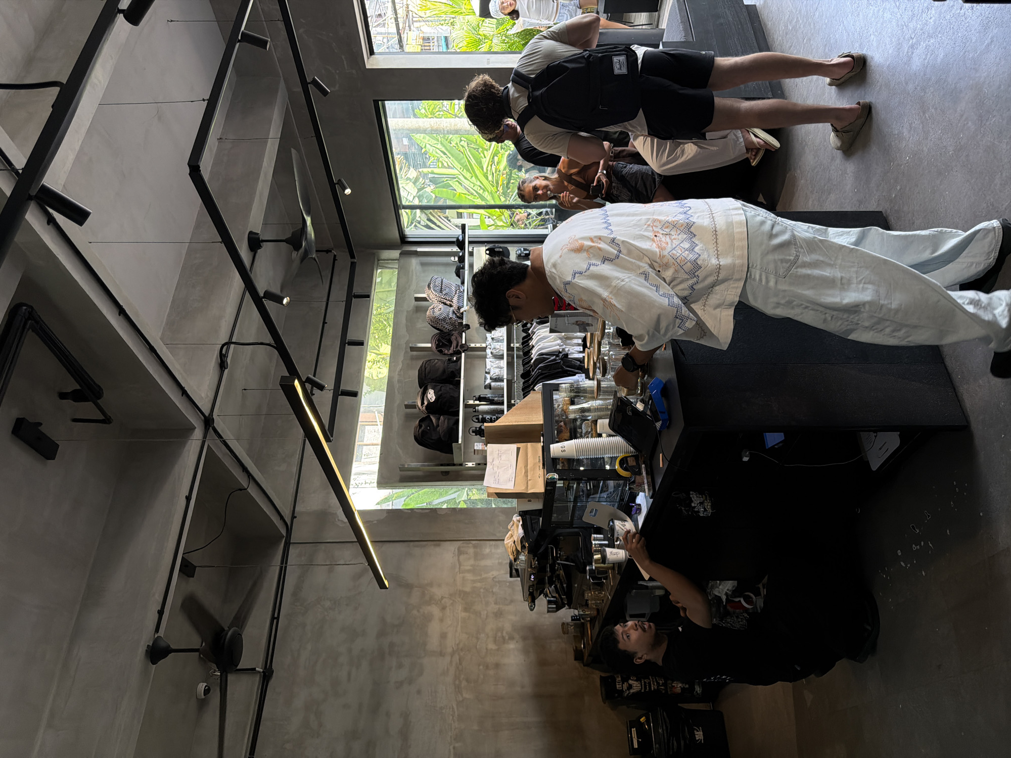 In a shop in North Kuta, Indonesia, several people are present. A person wearing a white and blue patterned shirt and light blue pants stands behind a dark counter, seemingly interacting with something on it. Another person in a black shirt is behind the counter as well. Two individuals, one wearing a backpack, are visible near a window. Shelves stocked with items are positioned against the wall. Overhead, a network of black metal beams supports lights and fans. The walls are a muted gray, and natural light streams in through the windows.