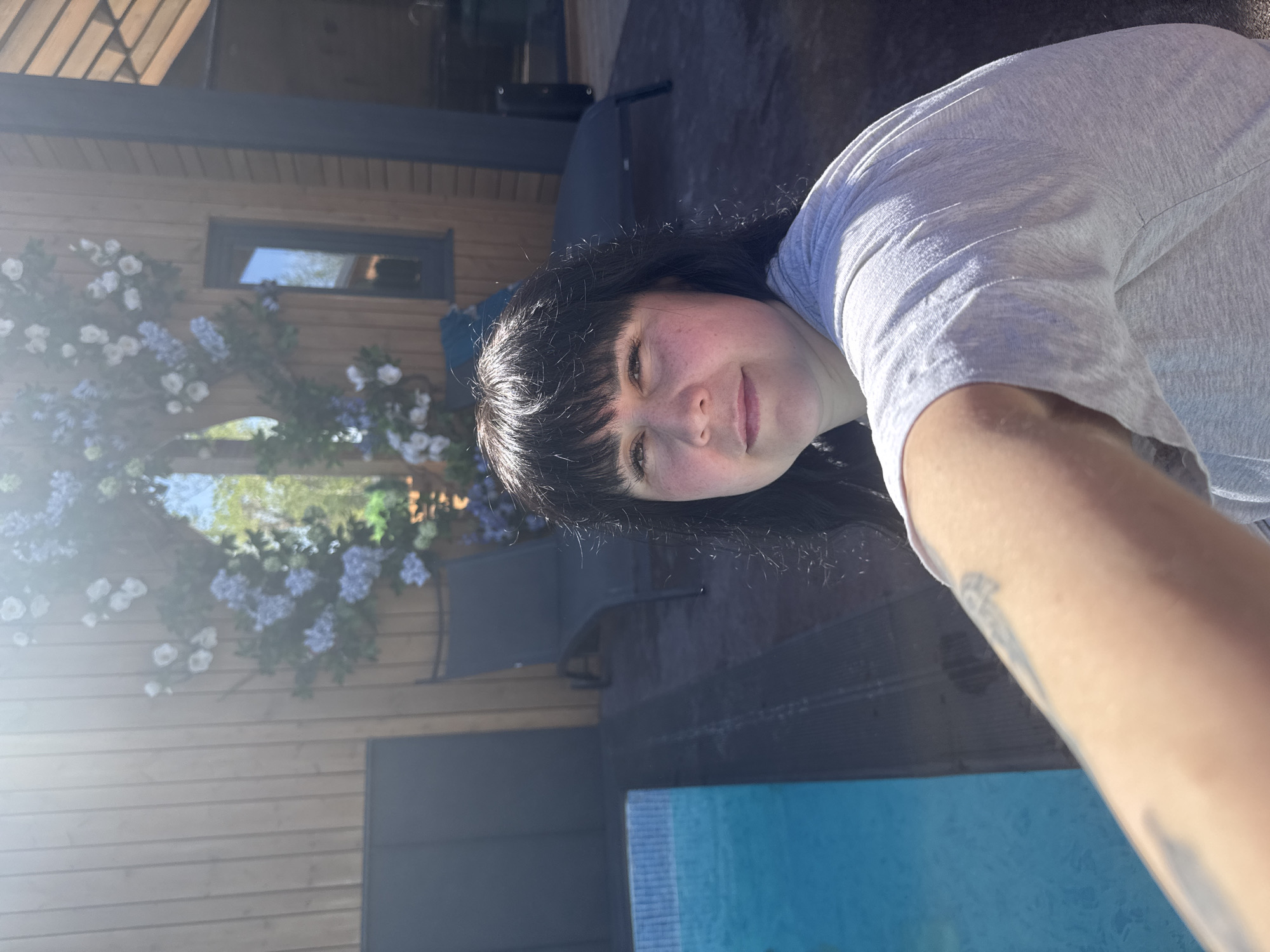 A selfie taken outdoors shows a woman with dark hair and bangs wearing a light gray t-shirt. A portion of a blue swimming pool is visible in the lower part of the frame. Behind her, there is a building with wood siding and a window. The building is decorated with white and blue flowers. There are chairs and a table visible in the background. The location is not identifiable as being in Izhevsk, Russia.