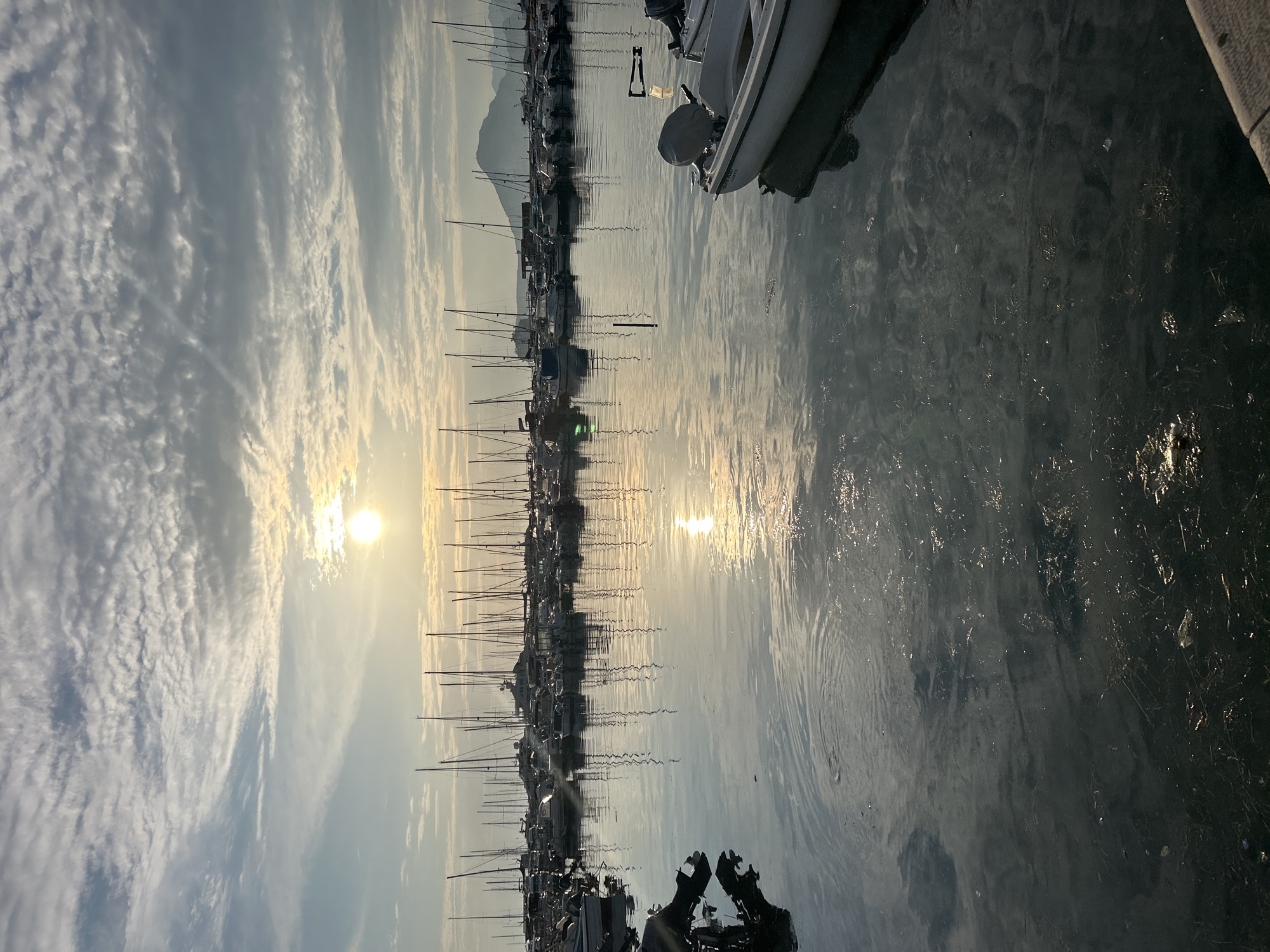 The image depicts a serene marina scene at sunrise or sunset, with the sun low in the sky and casting a golden glow on the water and clouds. The sky is filled with wispy, scattered clouds, creating a dramatic backdrop. A line of sailboats and yachts are docked, their masts stretching towards the sky, reflected in the calm water. The water itself is a mix of calm surfaces and rippling areas, with debris visible near the shore. A white boat with an outboard motor is prominently featured in the foreground, partially obscuring the view of the marina. In the background, mountains are faintly visible, suggesting a coastal or lakeside location. The overall mood is peaceful and tranquil, evoking a sense of calm and natural beauty. The image is oriented vertically, with the sky occupying the left side and the water and marina on the right. There are no visible people. No text is discernible in the image.
