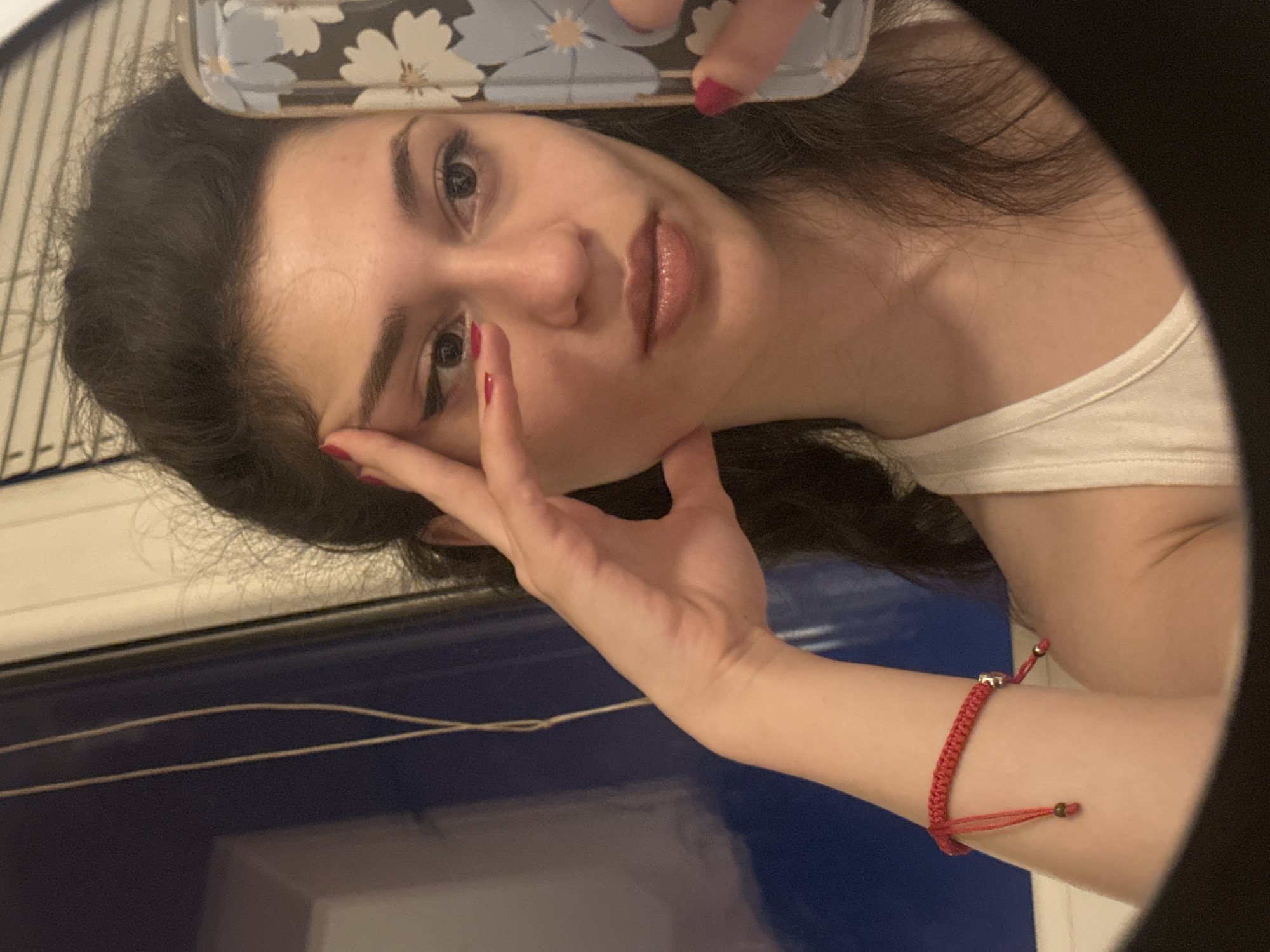 A young woman with dark, wavy hair is captured in a mirror selfie. She is wearing a white tank top and a red string bracelet on her left wrist. Her right hand is placed on her forehead, and her left hand holds a phone with a floral case, which is partially visible. She has on makeup, including eyeliner and lip gloss. The background is out of focus but appears to be an indoor setting with blinds and a dark blue surface. This image was taken in Sofia, Bulgaria.