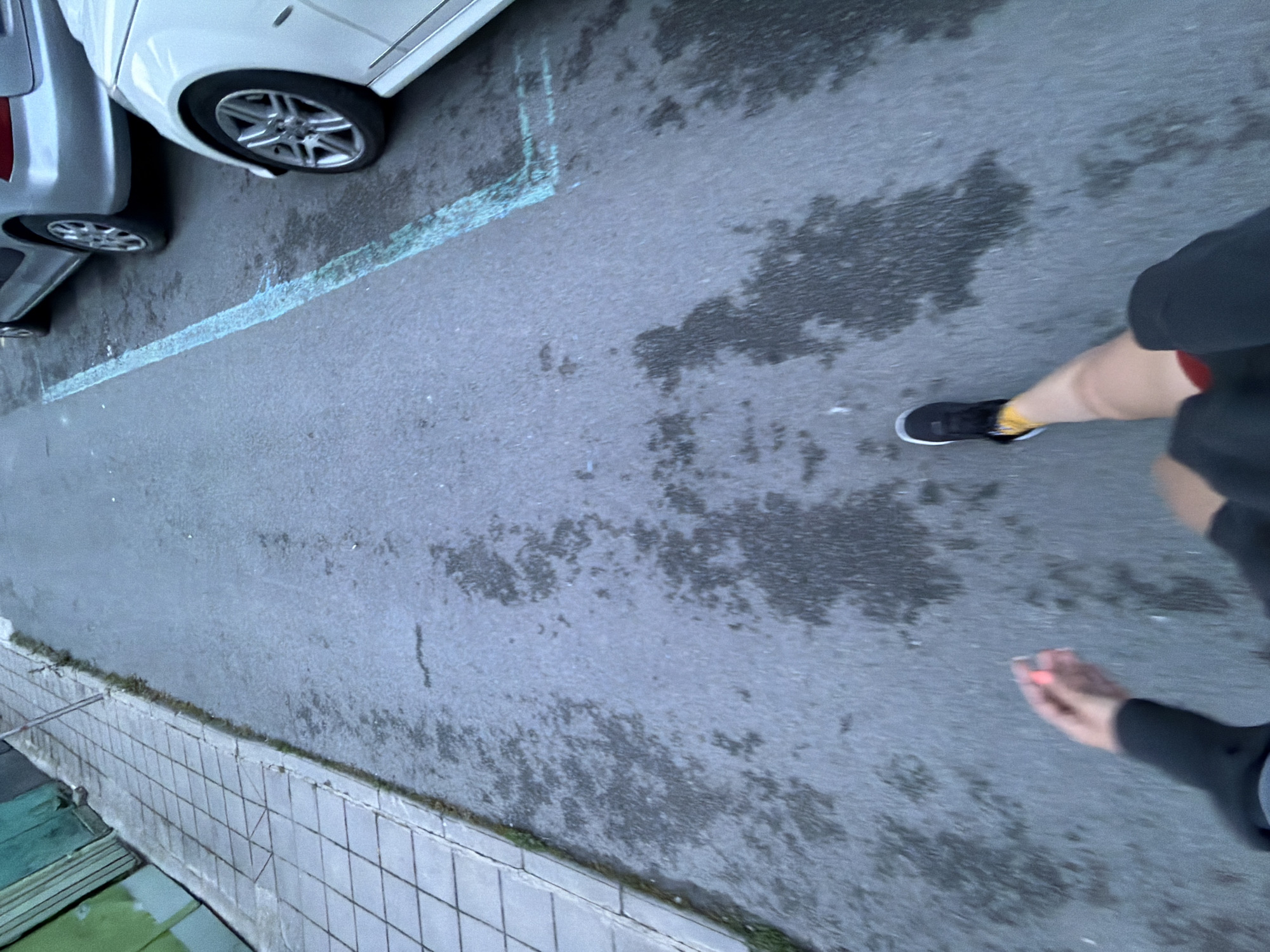 The image, captured from an elevated perspective, displays a paved outdoor area, likely a street or parking lot, in Burgas, Bulgaria.
On the left side of the frame, two cars are visible: a silver/grey vehicle and a white vehicle, both partially seen from their lower sections, including their wheels. A faded, light blue line is painted on the asphalt surface, possibly marking a parking boundary or an old road line. The asphalt also exhibits several dark, irregular patches, indicative of dampness or recent rainfall.
On the right side of the frame, a person is shown in motion, walking across the paved area. Their right leg is clearly visible, wearing a black athletic shoe with a white sole and bright yellow socks. Their left hand and forearm, covered by a dark sleeve, are also in view, holding a small, bright orange or red object.
Along the very bottom edge of the image, a tiled ledge or low wall, possibly part of a building or barrier, is visible. The lighting suggests it is daytime, though the damp pavement might imply overcast weather or recent precipitation. There is no discernible text present in the image.