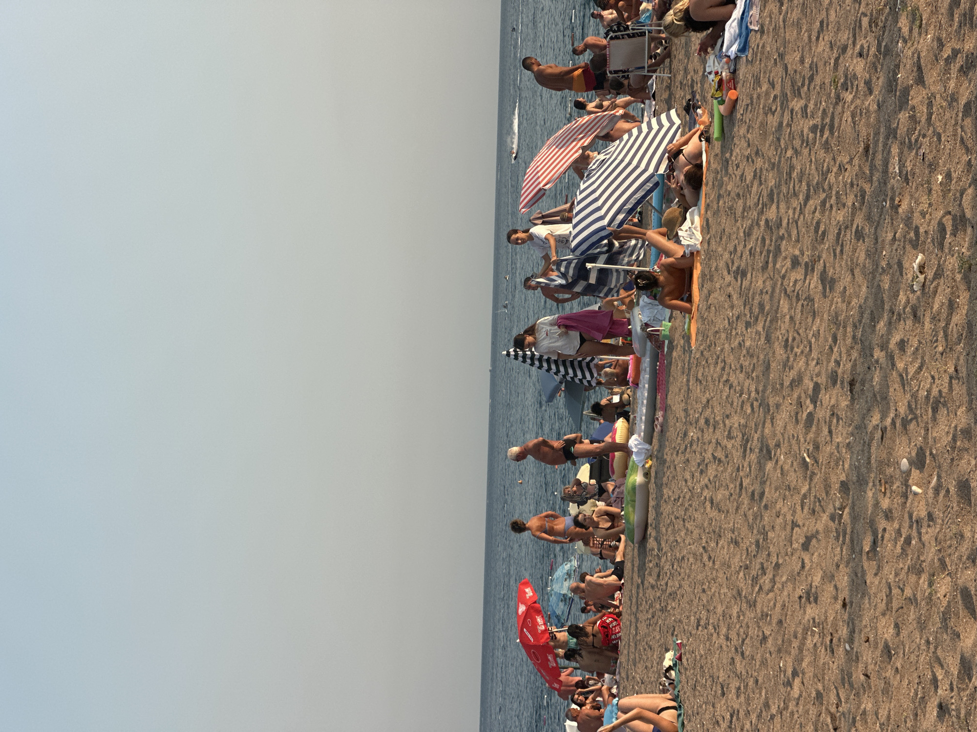 In Boreti, Montenegro, a crowded beach scene unfolds under a hazy sky. The sandy shore is dotted with numerous sunbathers, many relaxing on towels and inflatable mats. Beach umbrellas, both red and striped, offer shade to some. In the background, the calm blue sea stretches to the horizon, with a few distant jet skis indicating recreational activity. People of various ages are present, some wading in the shallows or strolling along the water's edge, while others are engaged in conversations or simply basking in the sun. The atmosphere appears relaxed and leisurely, characteristic of a popular summer destination.