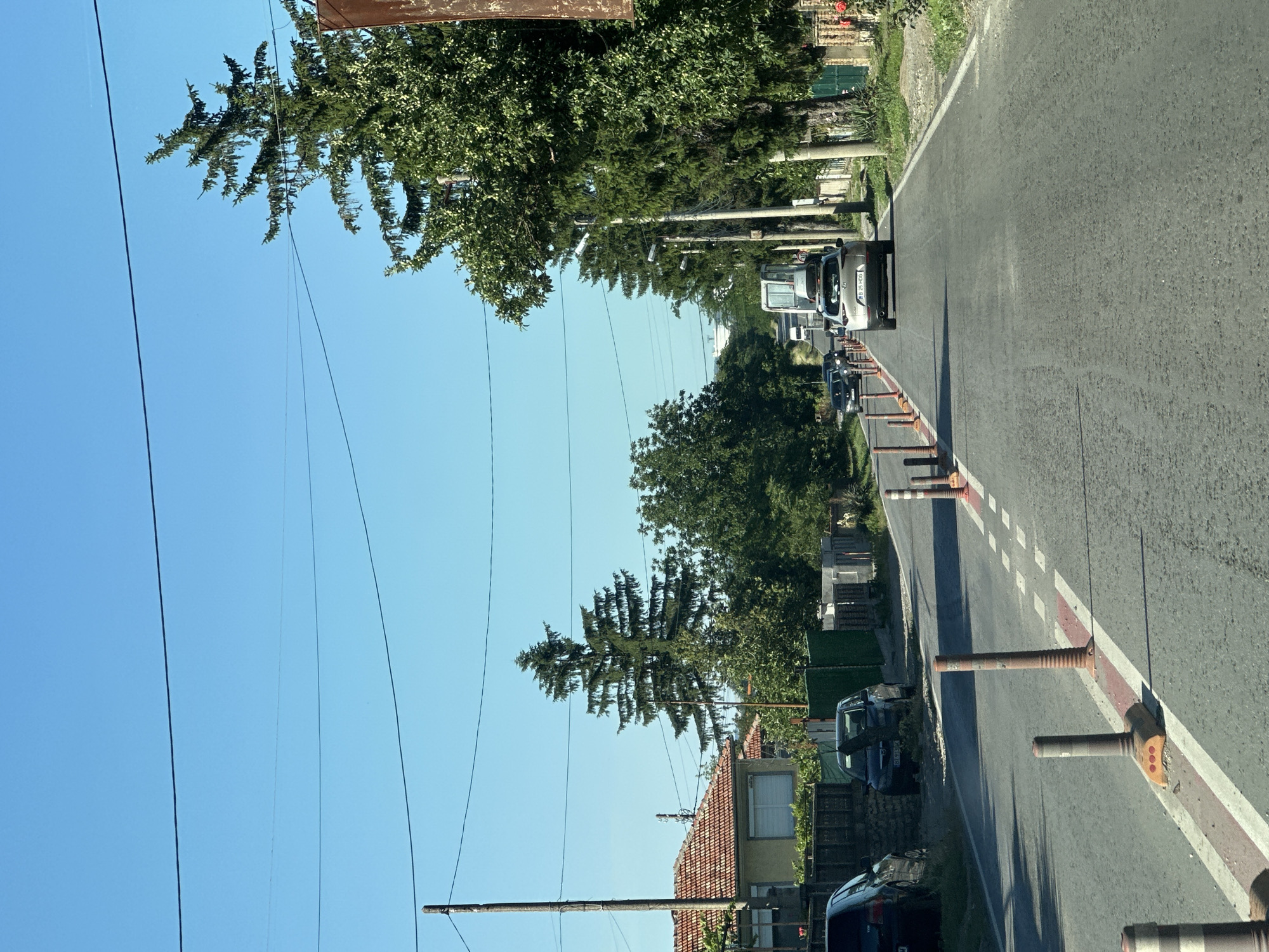 The image depicts a sun-drenched street scene in Samovodene, Bulgaria. The road, paved with asphalt, stretches into the distance, flanked by trees and residential buildings. A line of orange traffic cones with white reflective stripes divides the lanes. Several cars are visible on the road, including a silver SUV in the foreground. Buildings with red-tiled roofs and yellow facades line the left side of the street. Power lines crisscross the clear blue sky. The environment suggests a typical day in a Bulgarian village.