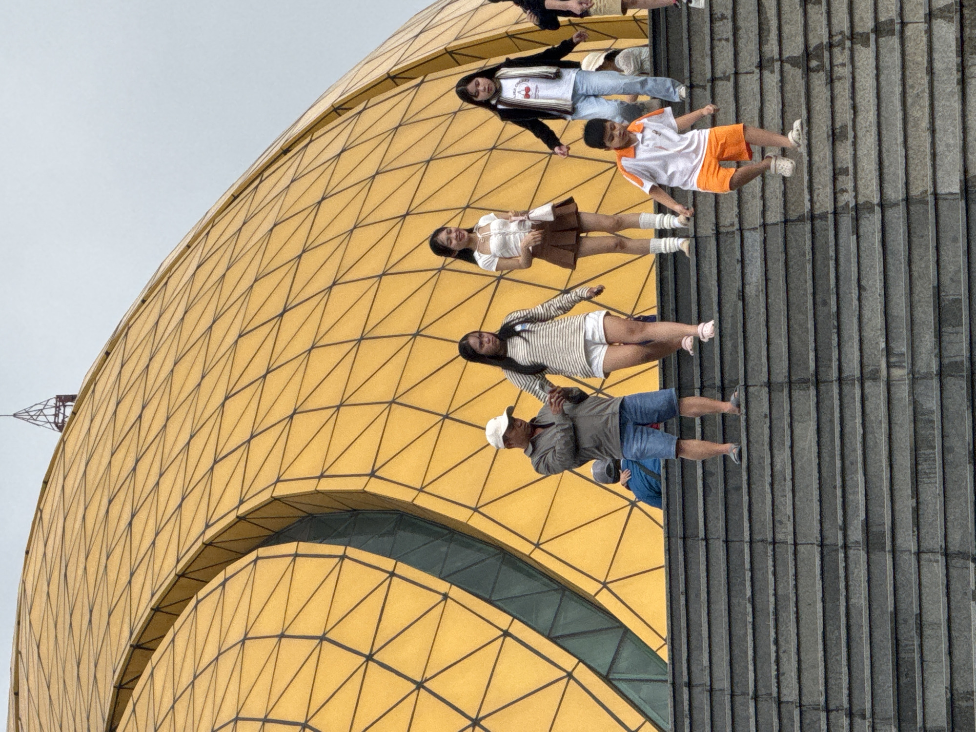 This image, taken in Da Lat, Vietnam, captures a group of people gathered on a wide staircase adjacent to a distinctive golden building under an overcast sky. The building, a prominent architectural feature, is characterized by its curved structure and a facade covered in a geometric, diamond-patterned grid of golden panels, strongly suggesting the Artichoke Bud building at Lam Vien Square. A tall, skeletal structure, possibly a crane or antenna, is faintly visible in the upper-left background, above the golden edifice.
Several individuals are positioned on the grey, multi-tiered steps. In the foreground, a man in a white cap, grey jacket, and blue shorts stands near a partially visible person in a blue shirt. Above them, a woman wears a striped long-sleeve top and white shorts. Further up the stairs, a smiling woman in a white top, brown skirt, and white knee-high socks poses. Beside her, a young boy in an orange and white polo shirt, orange shorts, and white shoes stands casually. Higher on the stairs, a woman is seen wearing a black jacket over a white t-shirt with the visible text "LIKE CHERRIES," paired with light blue jeans and white shoes. Another person is partially visible at the top of the stairs.
The people appear to be casually congregating or posing for photographs, indicative of tourists or visitors enjoying the unique architecture and public space. Their relaxed postures and smiles suggest a pleasant experience. The overcast sky provides soft, diffused lighting, creating an evenly lit scene without harsh shadows. The overall impression is that of a lively public space at a popular landmark.