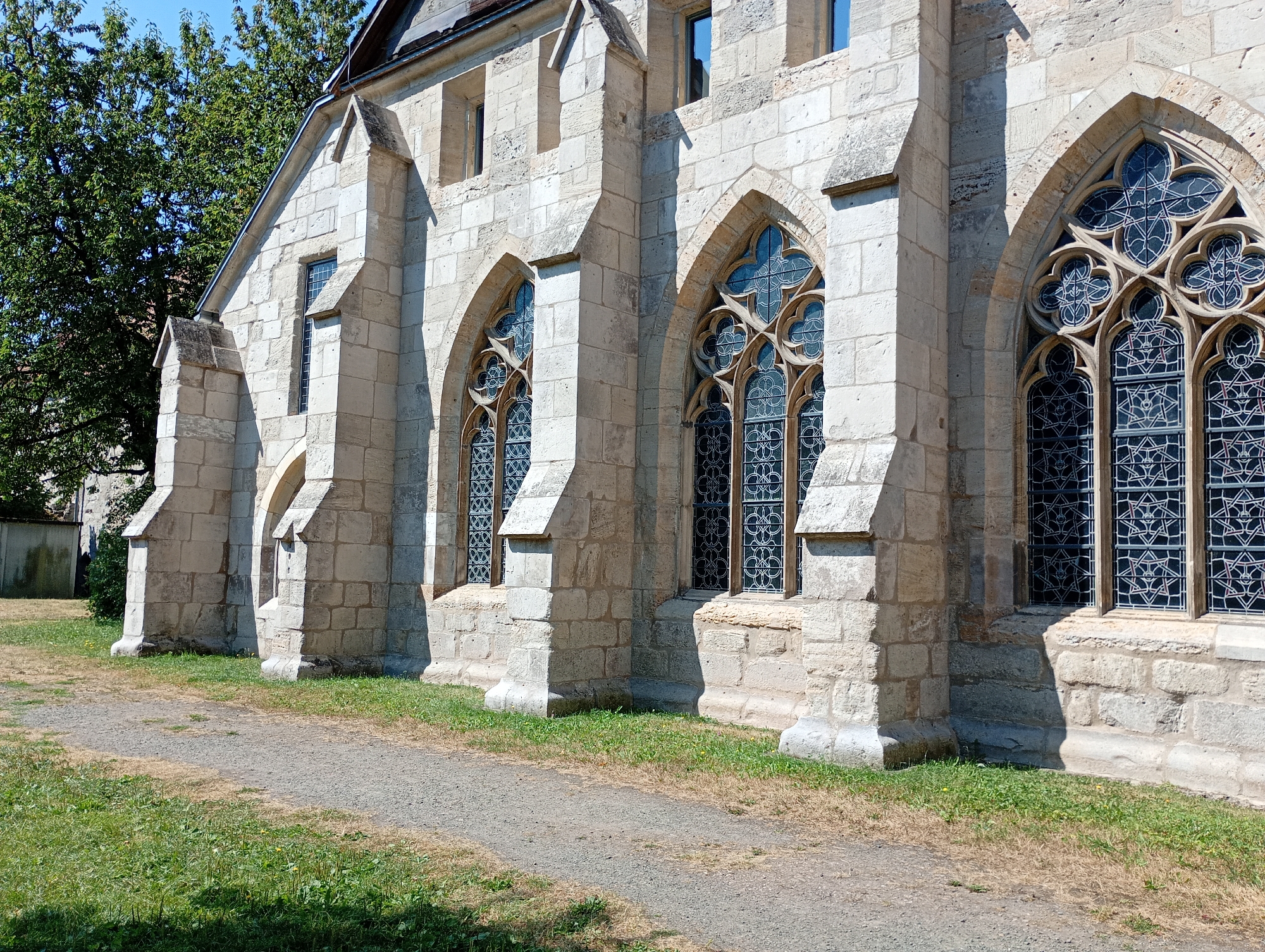 The image shows a close-up, low-angle view of the stone facade of a Gothic-style building, likely a church, in Walkenried, Germany. The building is constructed from large, light-colored stone blocks and features several arched windows with intricate tracery and dark glass. A gravel path runs in front of the building, bordered by a grassy area. Tall trees with green foliage are visible to the left of the building, casting shadows on the facade. The sky above is clear and blue. The scene is well-lit, suggesting it was taken on a sunny day. The architectural style and the presence of Gothic windows indicate it is likely part of the Walkenried Abbey complex, a significant historical site in the town. No people or animals are present in the image.