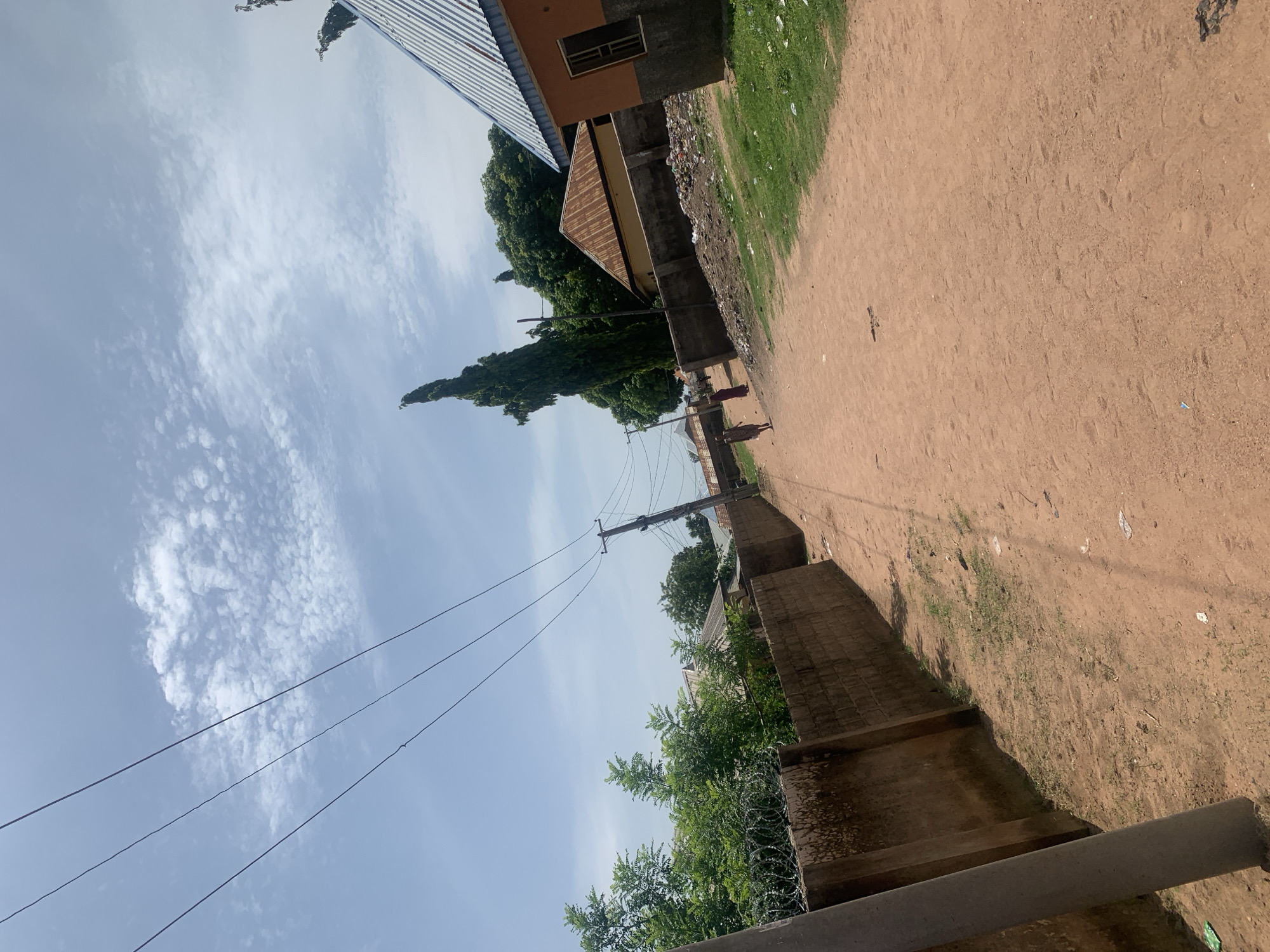 The image depicts a street scene in Jauro Yinu, Nigeria. A dusty, reddish-brown dirt road runs diagonally from the bottom right towards the upper left. On the left side of the road, a tall, gray concrete pole stands in the foreground, followed by a brick wall topped with barbed wire. Beyond the wall, lush green trees and some buildings with corrugated metal roofs are visible. Power lines stretch across the sky from a utility pole on the right side of the road. On the right side of the road, a building with an orange and gray facade and a corrugated metal roof is prominent. Further down the road, more buildings with various roof types and some piled-up refuse can be observed. Two individuals, appearing to be women dressed in traditional clothing, are walking away from the camera on the dirt road. The sky is a pale blue with scattered white, fluffy clouds. The overall environment suggests a semi-urban or rural setting.