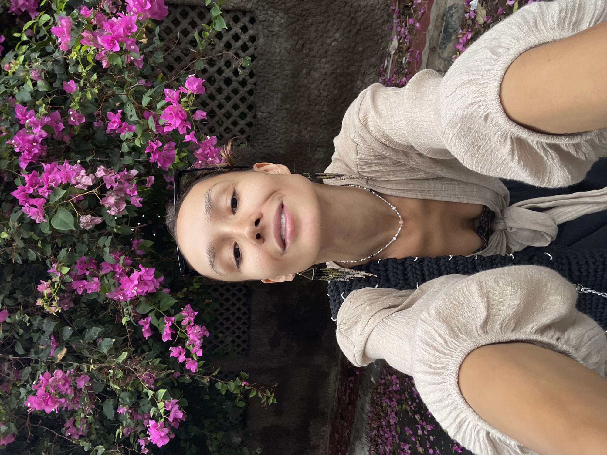 A woman smiles in an outdoor selfie taken in Phu Thuy Ward, Vietnam. A cascading bougainvillea plant, with vibrant pink flowers, serves as the backdrop to her left. She wears a light-colored blouse with voluminous sleeves, and a necklace. A dark, textured bag hangs from her shoulder. The background includes a wall with a lattice pattern and a glimpse of the street covered in fallen flowers.