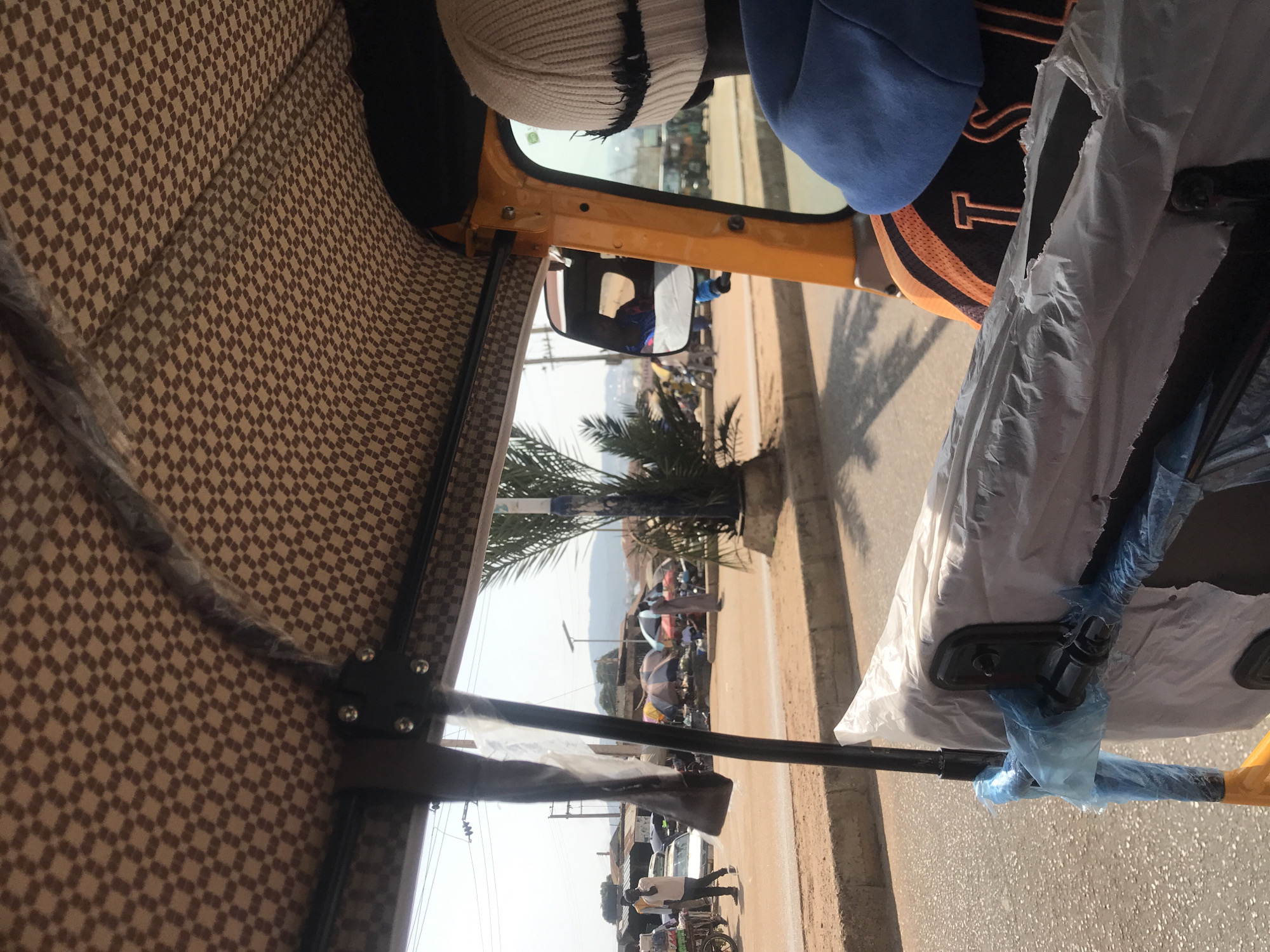 The image provides a view from inside an auto-rickshaw, likely in the city of Jos, Nigeria, looking outwards. The interior of the auto-rickshaw features a patterned brown fabric lining the roof. Outside, a dusty road is visible, lined with shops and market stalls. Several people are present, including a man walking on the left, a person standing near a stall in the distance, and others scattered around the market area. Various vehicles, including cars and other auto-rickshaws, are also present. The environment appears to be a busy marketplace on a bright, sunny day, with a slightly hazy sky suggesting a warm climate. The presence of market umbrellas and people engaged in commerce points to a typical urban market scene in Nigeria.