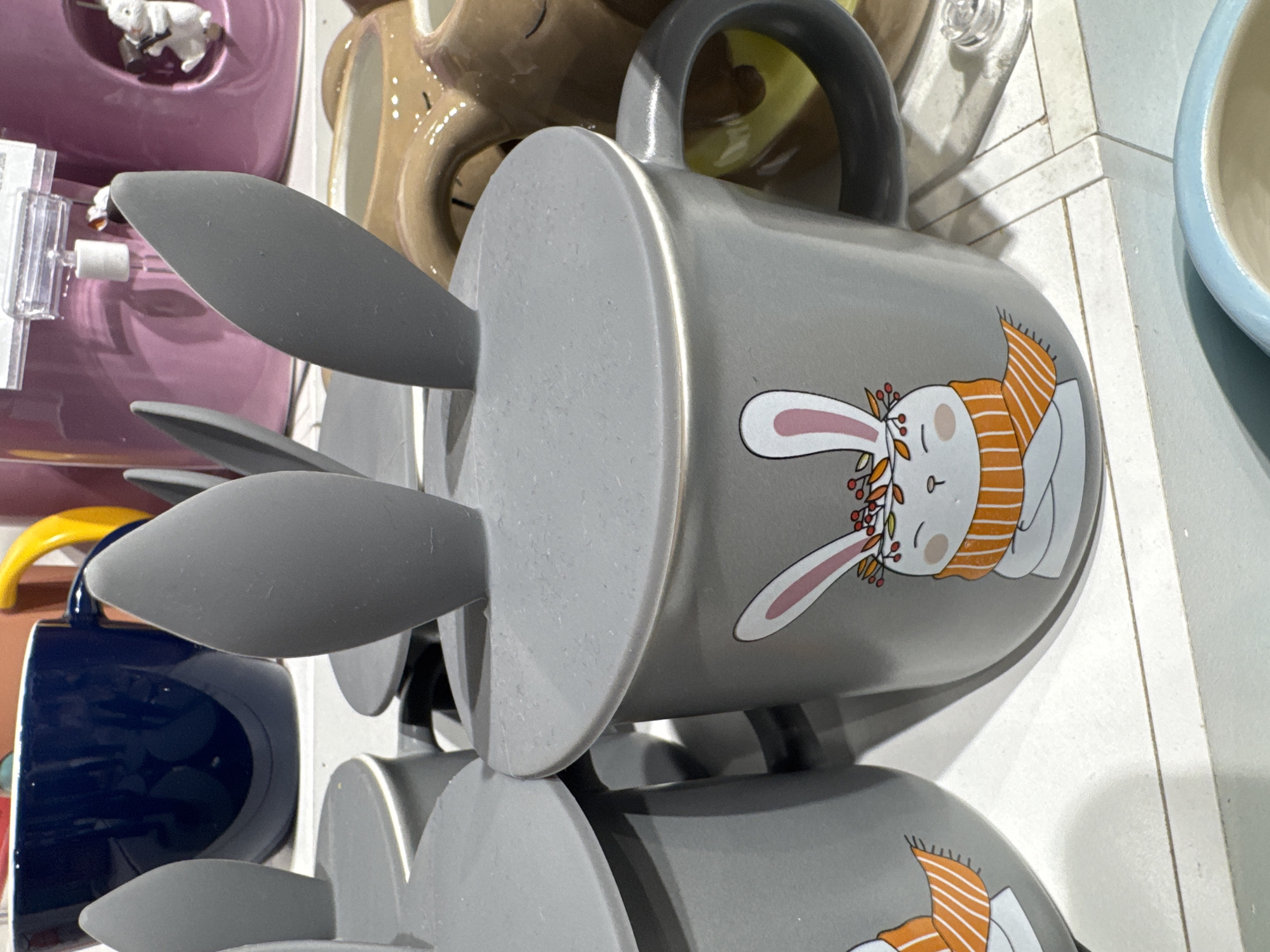The image displays a close-up view of several mugs with a whimsical design, focusing on one prominent gray mug. This mug is adorned with a cartoon illustration of a rabbit wearing a festive orange and white striped scarf and a crown of leaves and berries. The mug also features a grey lid with two protruding, soft, ear-like shapes, resembling bunny ears. In the background, other mugs are visible, including a dark blue mug, a pink mug with a small white figurine, and a brown mug shaped like a bear. The scene appears to be a retail display, likely in a store, with shelves or surfaces holding the merchandise. The lighting is bright, suggesting an indoor environment. The specific location in Ufa, Russia, cannot be determined from the visual information provided in the image.