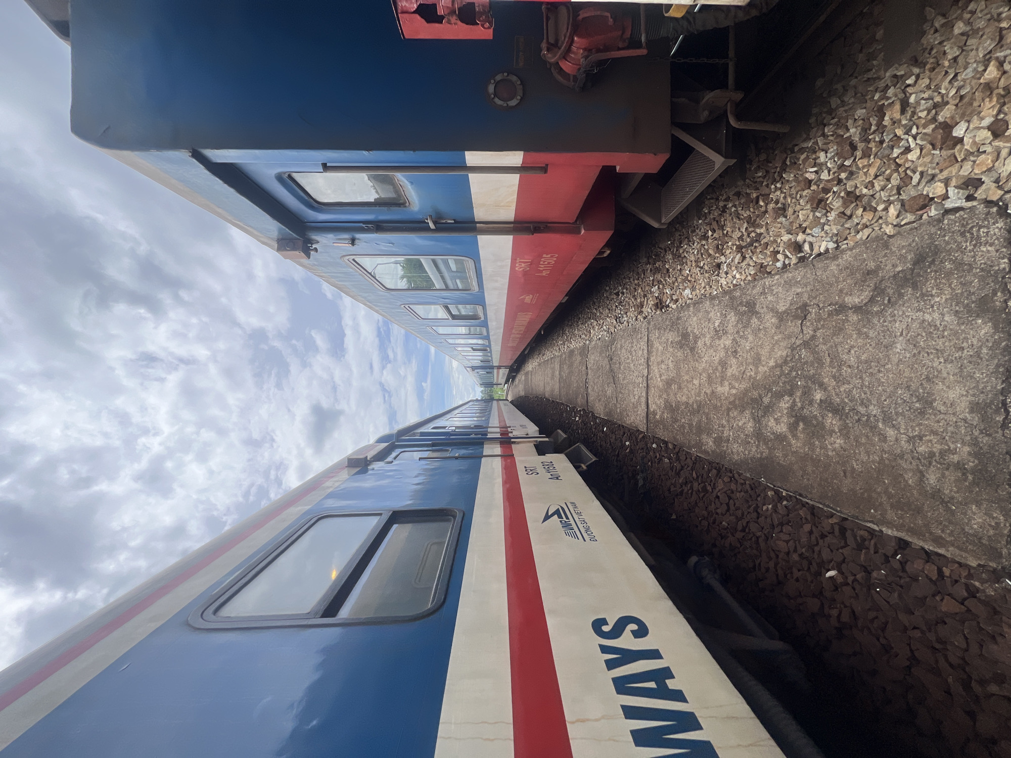 Two blue and white trains with red stripes are parked side-by-side on train tracks in Huế, Vietnam. The trains are positioned at an angle, with the perspective drawing the viewer's eye down the narrow space between them. The sky above is partly cloudy. The train on the left has "WAYS" visible in large blue letters on its side, followed by smaller text that includes "VIETNAM RAILWAYS" and a logo. The train on the right has "SRT AN11505" printed on its side. The ground between the trains is a concrete path bordered by ballast stones. The overall scene suggests a train station or depot.