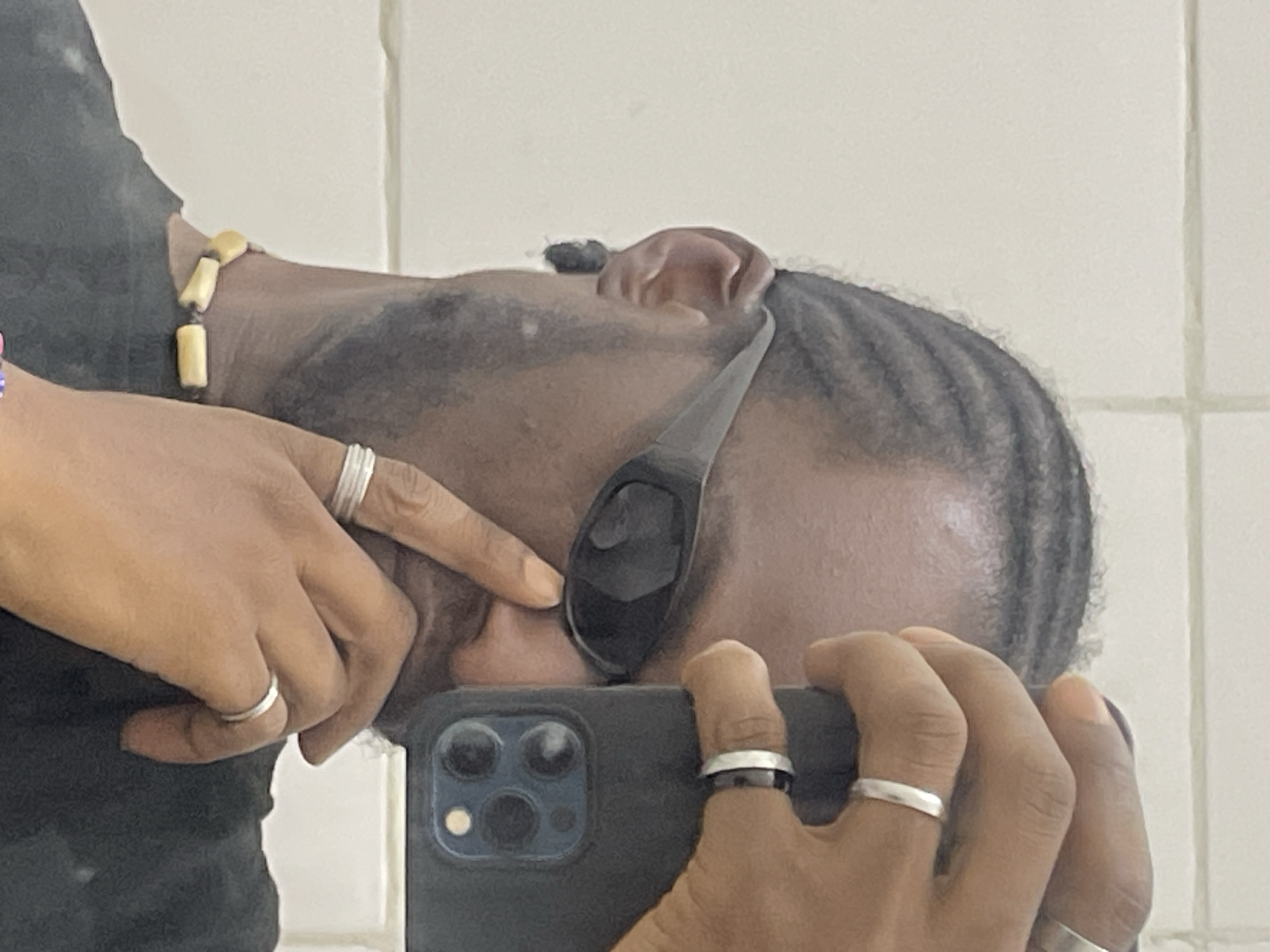 The image captures a dark-skinned individual taking a mirror selfie in Jos, Nigeria. The subject's head is angled, revealing cornrows pulled back on the scalp and a faint beard/stubble along the jawline. He is wearing dark, rectangular wrap-around sunglasses and a dark-colored garment, likely a shirt. A necklace with light-colored, irregular beads is visible around his neck. Multiple silver-colored rings adorn his fingers: one simple band and one wider band with a dark inlay on his right hand (which holds the smartphone), and two simple bands on his left hand. The left index finger is positioned near the bridge of the sunglasses. The smartphone, encased in a dark cover, partially obscures the subject's lower face. Its rear triple-lens camera module and flash are visible. The background consists of a light-colored, tiled wall with visible horizontal and vertical grout lines, indicative of an indoor setting.