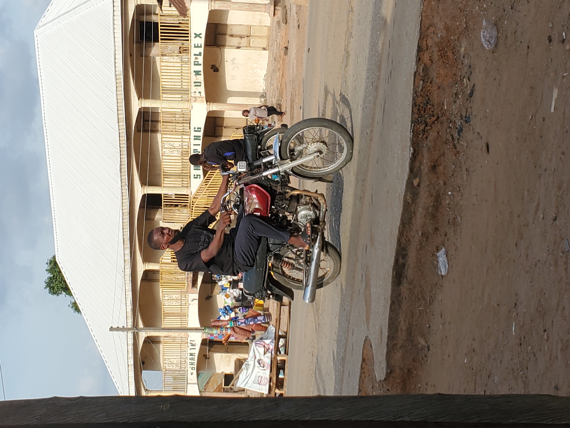 A street-level photo captures a man seated on a red motorcycle in what appears to be a commercial area in Mararaba, Nigeria. The man, wearing a black t-shirt with "No Thanks" written on it and dark pants, is positioned to the left of the frame, looking down towards the motorcycle's controls or seat. His feet are in sandals. Behind him, a second man in a dark blue and black top stands next to another motorcycle, partially visible.
The scene features a paved road with a dirt shoulder on the right, strewn with some litter. In the background, a large, multi-story building dominates, featuring a white corrugated metal roof and multiple balconies with yellow metal railings. Vertically inscribed on the building's facade are the words "SHOPPING COMPLEX," with "SHAM IK" partially visible below, likely part of a name. The bright daylight and clear sky suggest it is daytime, casting visible shadows. The overall impression is a typical street environment in an urban or suburban setting.