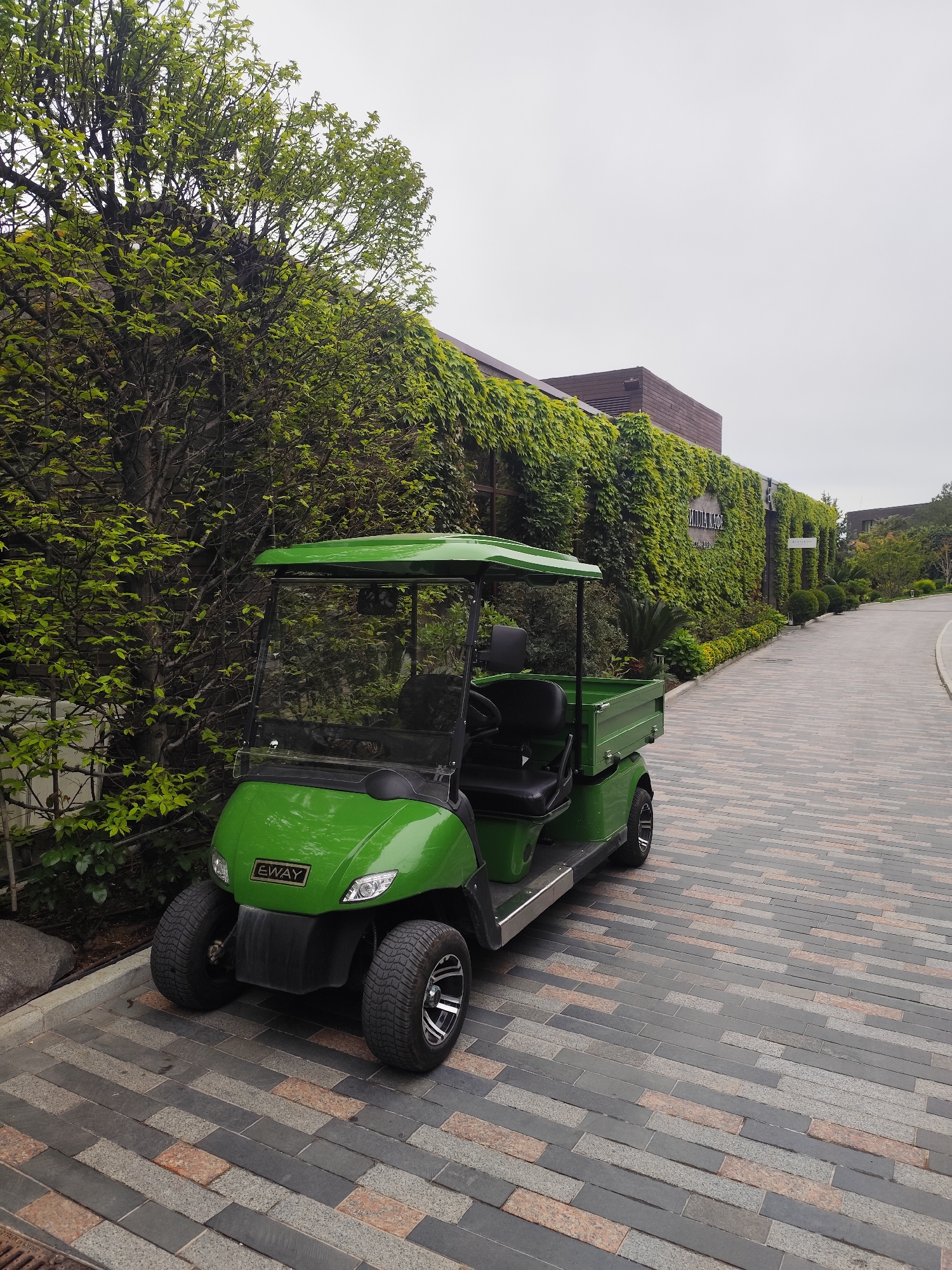 The image shows a green utility vehicle parked on a brick paved road, next to a wall covered in green foliage. The vehicle has "EWAY" written on the front. The road curves into the distance towards a building also covered in foliage. The scene appears to be located in Симеизский поселковый совет, Ukraine, given the presence of Cyrillic lettering on the building in the background. There are no people visible in the image. The overall environment is lush with greenery.