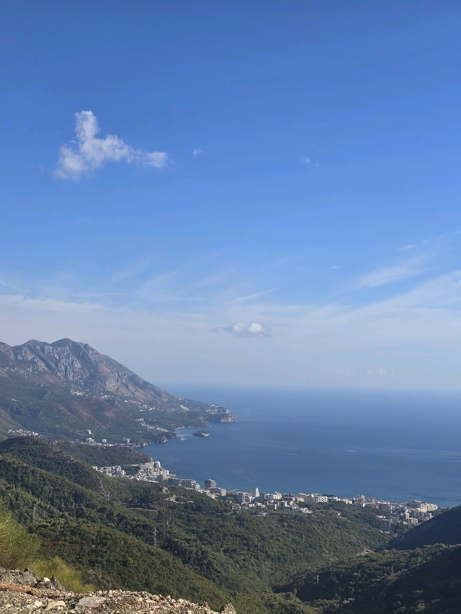 This is a vertical, medium-shot, eye-level, natural light, wide-angle photograph taken from a high vantage point looking down on a coastal city nestled between verdant mountains and the Adriatic Sea. The sky is a clear, vibrant blue with scattered wispy white clouds. The ocean, a deep cerulean, stretches to the horizon, with subtle white ripples indicating distant activity and a few small boats.
In the mid-ground, a densely populated coastal town, identified as being in the vicinity of Lapčići, Montenegro, sprawls along the shoreline. Buildings of varying heights and architectural styles, predominantly white and light-colored, are clustered together, forming a stark contrast against the lush green of the surrounding hillsides. The terrain slopes dramatically from the mountains down to the sea, with the town occupying the gentler slopes and the flat coastal strip.
The foreground is dominated by a steep, rocky incline covered in sparse vegetation, including dry grasses and small shrubs, suggesting an arid or semi-arid environment. The rugged texture of the rocks and the shadows they cast add depth and a sense of ruggedness to the scene. In the distance, a range of imposing, rugged mountains rises, their peaks etched against the sky, partially obscured by a haze. The overall impression is one of a picturesque coastal landscape, showcasing the dramatic interplay between mountains, sea, and human settlement. There are no people visible in the image.