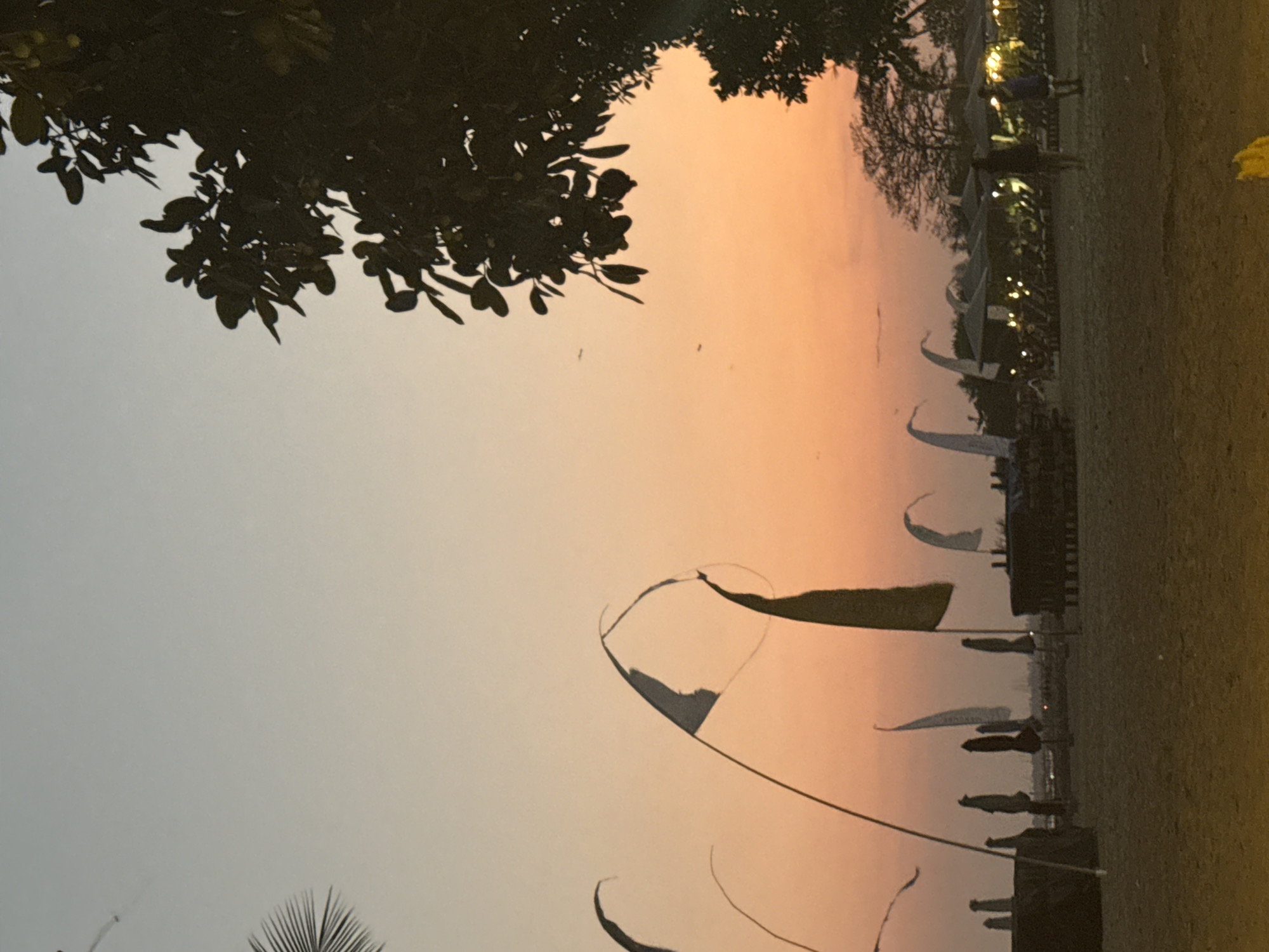 The image depicts a beach scene at dusk in Denpasar, Indonesia, likely during a sunset. The sky exhibits a gradient of soft colors, transitioning from a pale blue at the top to warm orange and pink hues closer to the horizon. In the foreground, a sandy beach is visible, with several beach umbrellas and lounge chairs set up. Small flags or pennants are fluttering on poles, adding a touch of movement to the scene. A silhouetted tree with dense foliage is present on the right side of the frame, casting a shadow. In the background, the lights of what appear to be beachside establishments are illuminated, creating a festive atmosphere. A few figures are walking on the beach, their forms indistinct in the low light. The overall impression is one of a tranquil evening by the sea.