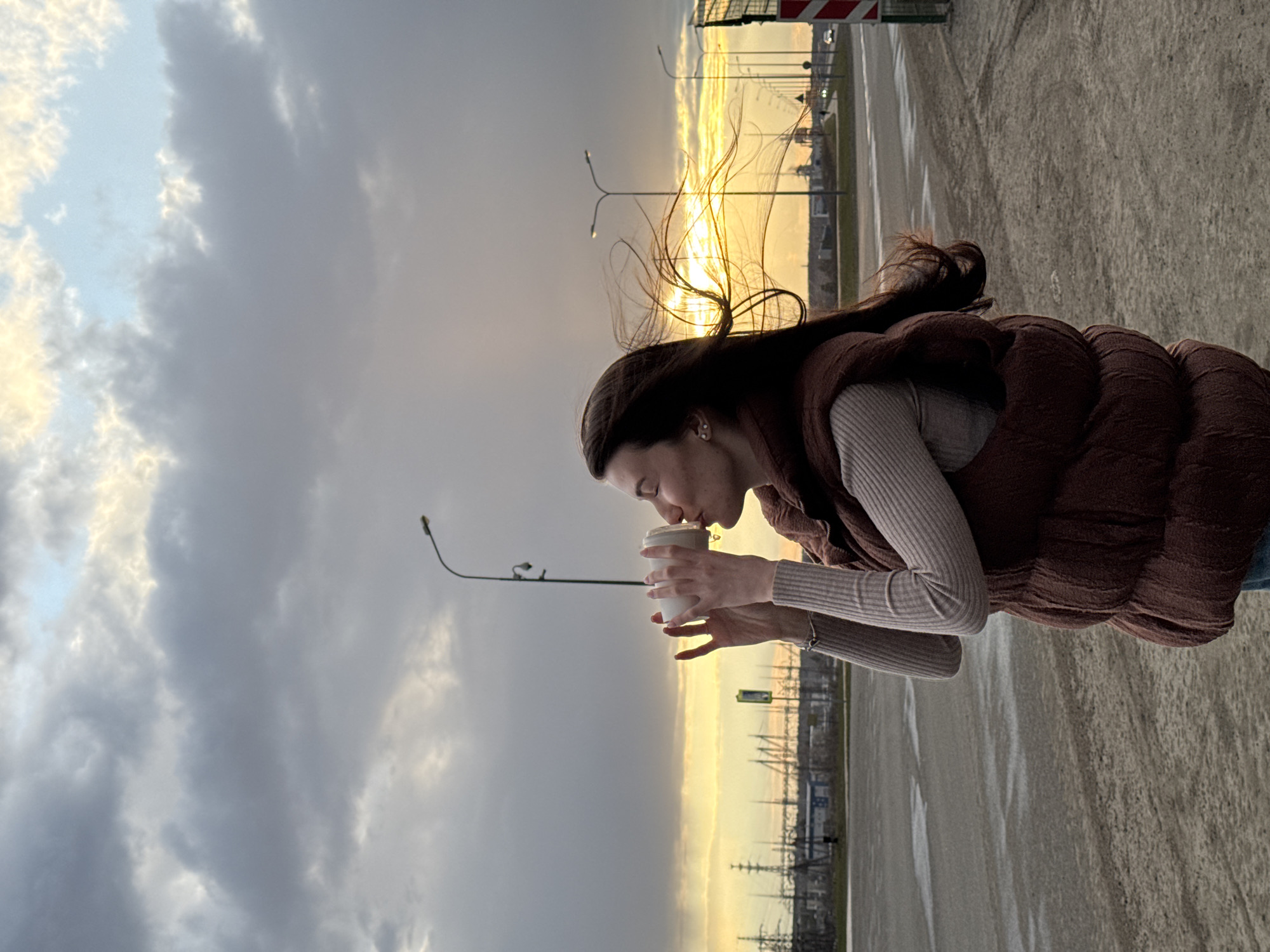 A young woman stands outdoors, holding a white disposable cup to her mouth as if drinking or smelling its contents. Her long, dark hair is swept back by the wind, with strands blowing towards the golden sunset horizon. She is dressed in a dark brown puffy vest over a light-colored, ribbed long-sleeve shirt. A small, shiny earring is visible in her right ear.
The scene depicts a wide, paved area, possibly a road or a large parking lot, extending into the distance. The background is dominated by a dramatic sky: dark, heavy grey clouds cover the upper portion, contrasting sharply with a vibrant golden glow from the setting or rising sun on the horizon. Silhouetted against this bright light are several streetlights and distant structures that appear to be industrial or utility-related, possibly power lines or factory buildings. A red and white striped barrier or sign is partially visible on the far right. The ground is a textured grey, suggesting asphalt or concrete, with some lighter, smoother patches that could be puddles or wet reflections. The overall impression is of a cool, windy evening in Chaikovsky, Russia, with a moment of quiet reflection. No visible text is present in the image.