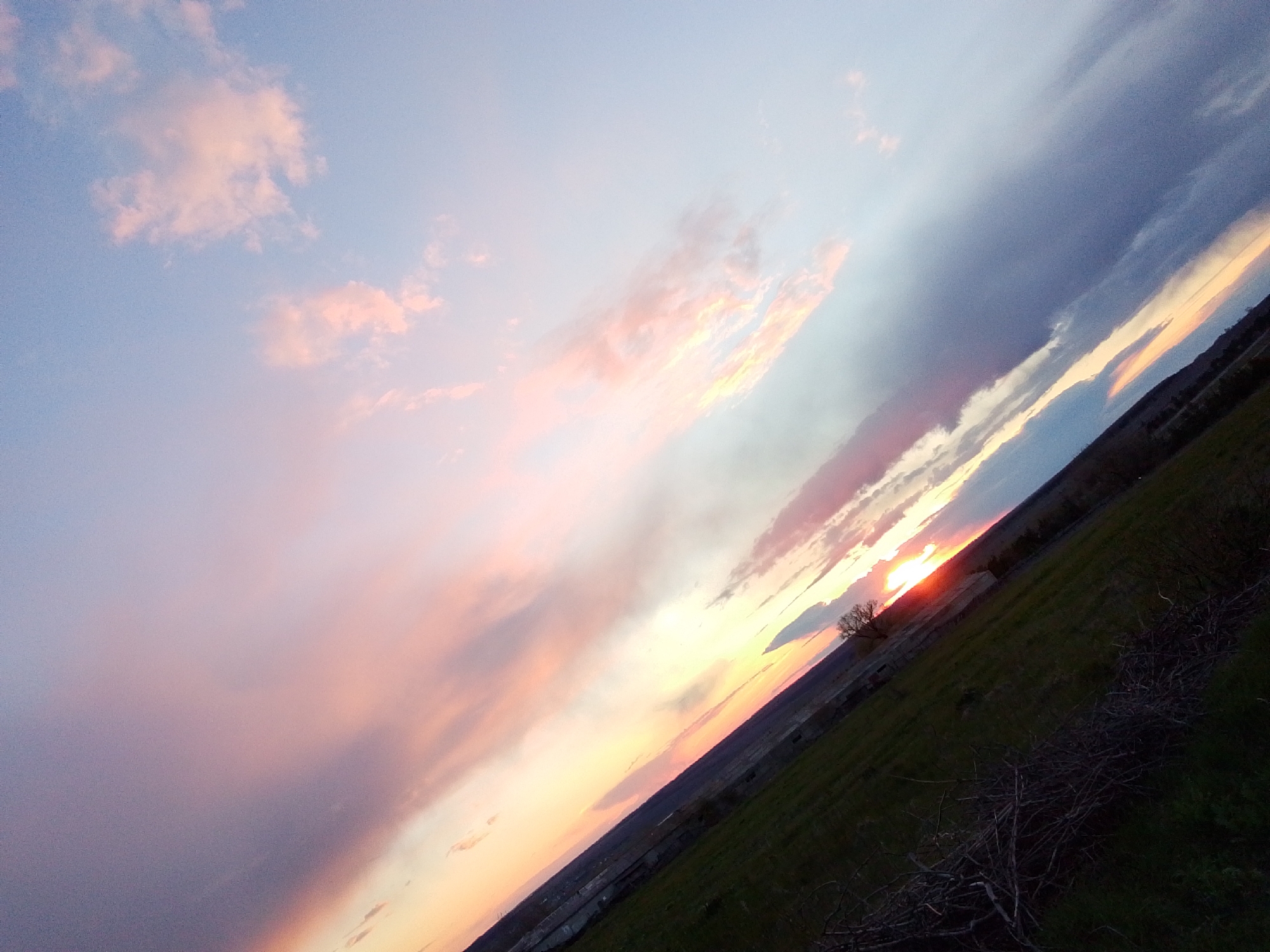 The image captures a vibrant sunset or sunrise over a rural landscape in Khlevnoye, Russia, characterized by a striking diagonal composition. The horizon line runs from the lower right to the upper left, tilting the entire scene.
The sky dominates the upper two-thirds of the image, showcasing a spectacular array of colors. On the far left and top, the sky transitions from a soft, pale blue to delicate pink and purple hues, with scattered, diffuse clouds catching the light. Towards the center and right, the colors intensify into bright oranges, yellows, and deeper pinks, concentrated around the setting or rising sun. The sun itself is a brilliant, glowing orb, partially obscured by distant land features and clouds, casting a warm light across the lower part of the sky. Adjacent to these bright areas, darker, more stratified clouds, ranging from deep grey to purplish-blue, stretch across the sky.
The lower third of the image depicts a dark, grassy field that appears to be a foreground. This field is mostly shadowed, with visible patches of dark green grass and several piles of dried branches or brushwood towards the bottom right. In the mid-ground, a distant, dark silhouette of land or hills stretches along the horizon line. A lone, bare tree stands silhouetted against the bright sun, its branches reaching upwards.
There are no people or discernible activities taking place. The overall impression is one of natural beauty and tranquility at the transition of day and night. No visible text is present in the image.