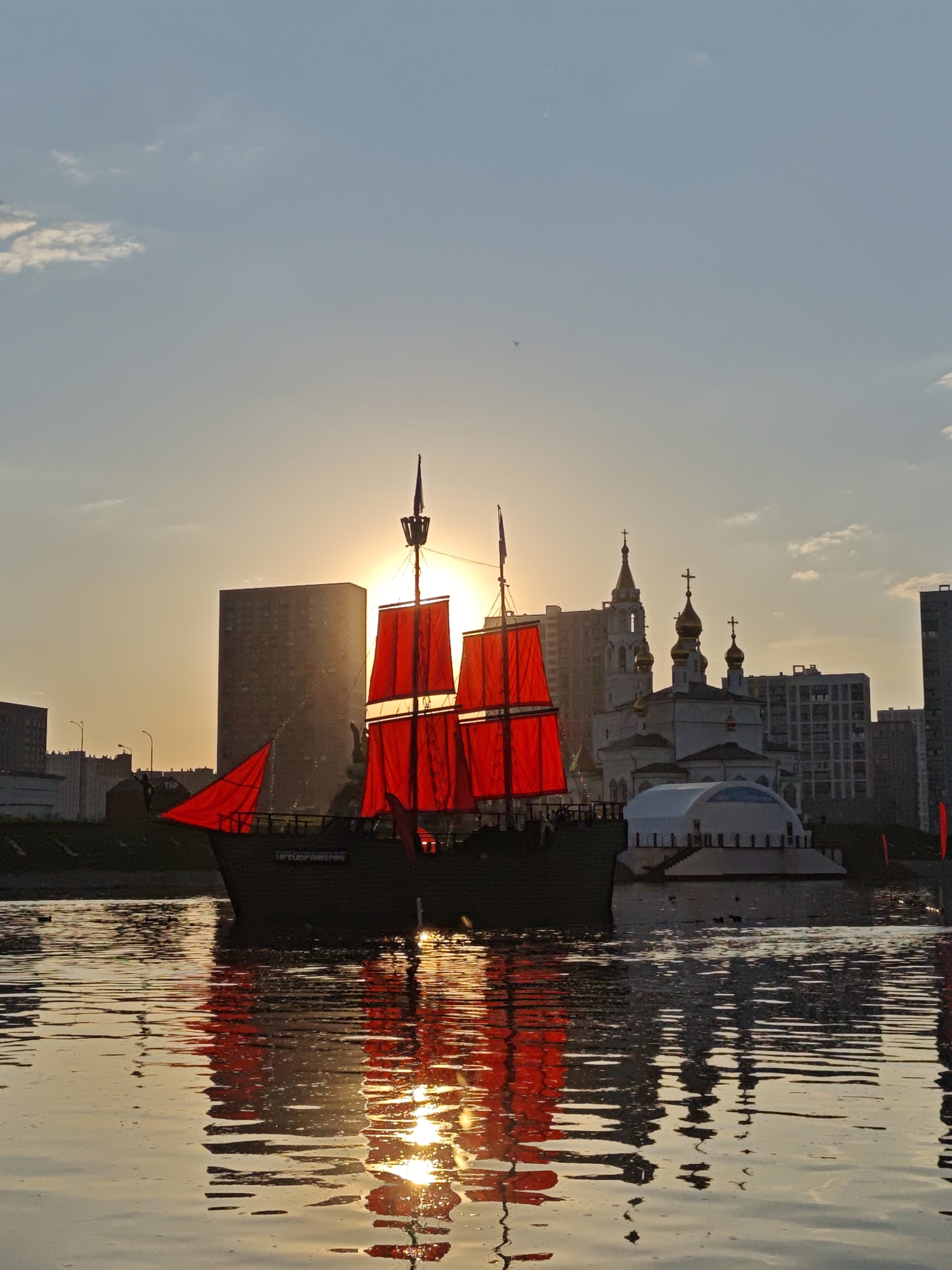 A low-angle shot showcases a body of water reflecting the setting sun, with a large replica ship featuring red sails in the foreground. The ship is docked near the city's waterfront, with a silhouette of a church featuring several domes and a building with modern architecture in the background. The setting sun is behind the ship's masts, creating a backlit effect. The lighting suggests either dawn or dusk. The location is Yekaterinburg, Russia.