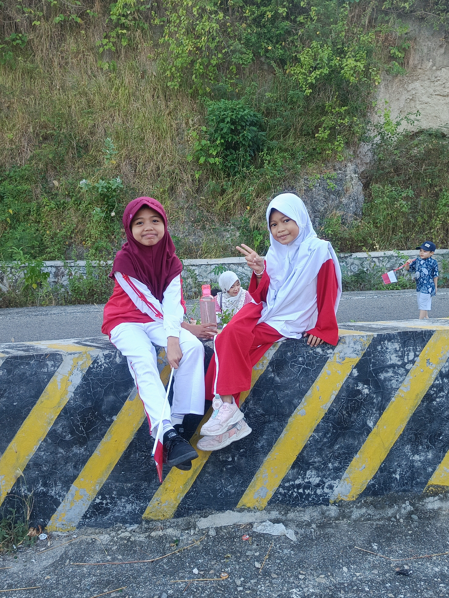 Two young girls in red and white hijabs and matching outfits sit on a concrete barrier painted with black and yellow diagonal stripes. The girl on the left is wearing a maroon hijab and looking at the camera with a slight smile. The girl on the right is wearing a white hijab and giving a peace sign with her left hand, smiling at the camera. Behind them, a third child, a boy wearing a blue patterned shirt and a blue cap, stands holding a small Indonesian flag. The background is a rocky embankment covered with dry grass and green foliage. The ground in front of the barrier is covered with gravel and debris. The scene appears to be outdoors, possibly on a road or path. The image is taken from a slightly low angle, looking up at the girls. The lighting suggests it is daytime, likely in the afternoon due to the shadows. The overall impression is one of a casual outdoor gathering, possibly related to a local event or celebration given the Indonesian flag.