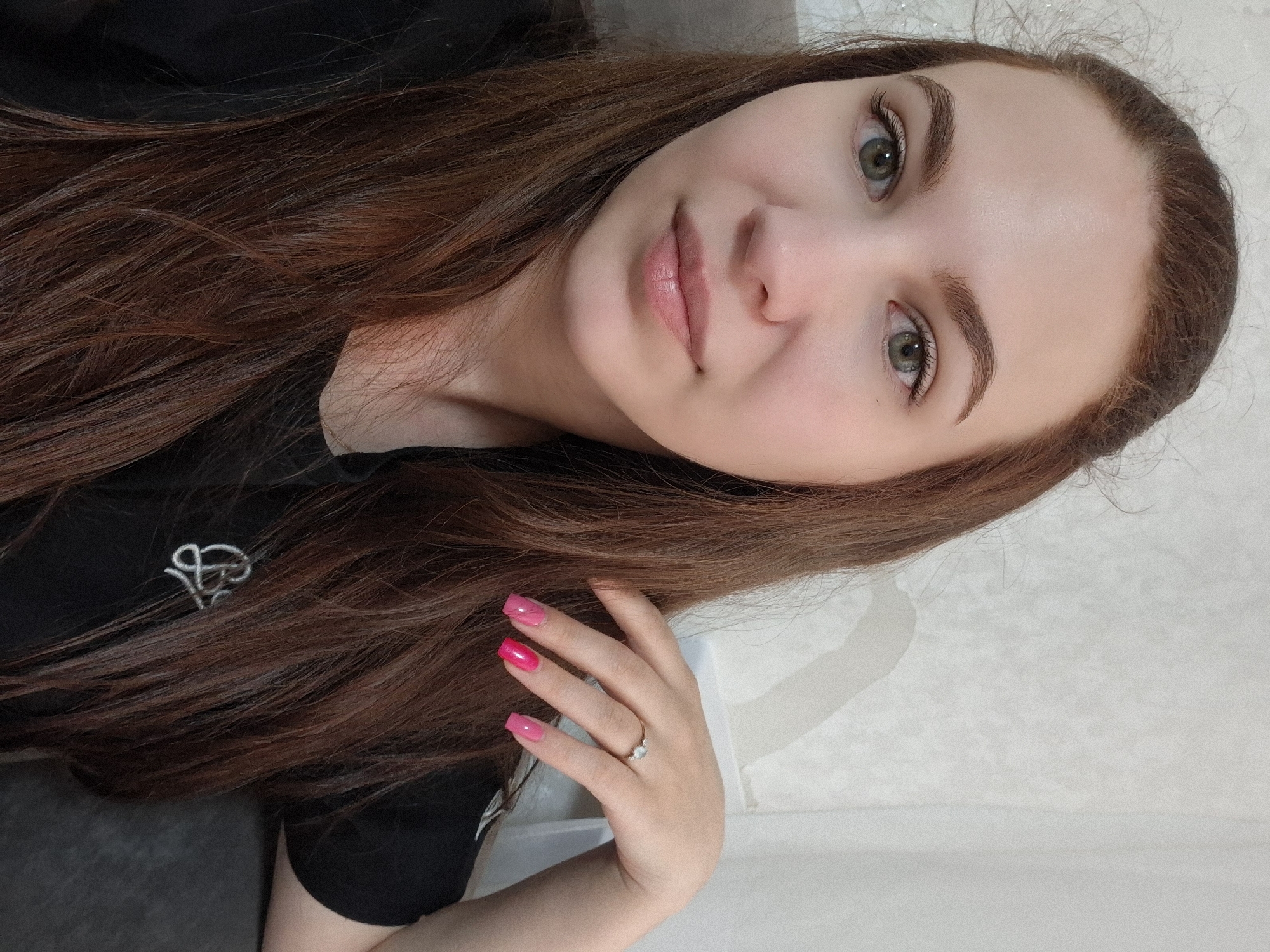 A selfie of a woman with auburn hair, green eyes, and fair skin. She is wearing a black top with a white logo on the left breast. Her nails are painted with a gradient of pink. She is wearing a ring on her ring finger. The background is a light-colored wall. The location is unspecified, and there is no information available to suggest the location is Chaikovsky, Russia.
