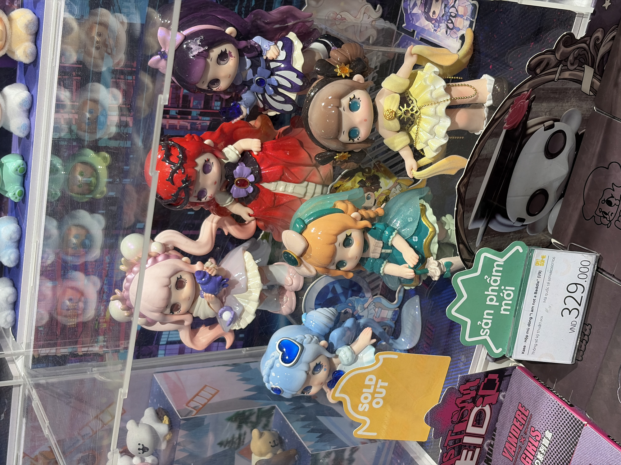 The image depicts a display case filled with "I am not a Baddie" collectible dolls, likely within a retail setting in Thủ Đức, Vietnam, as the price tag indicates Vietnamese Dong (VND). The dolls are arranged in rows, featuring various character designs with different color schemes (purple, red, pink, orange, and blue). A "SOLD OUT" sign is visible near the blue doll. A green sign reads "sản phẩm mới", indicating "new product". A price tag is also visible, stating "VND 329.000". Other plush toys are also on display in the left section of the display case. The display case has clear glass panels.