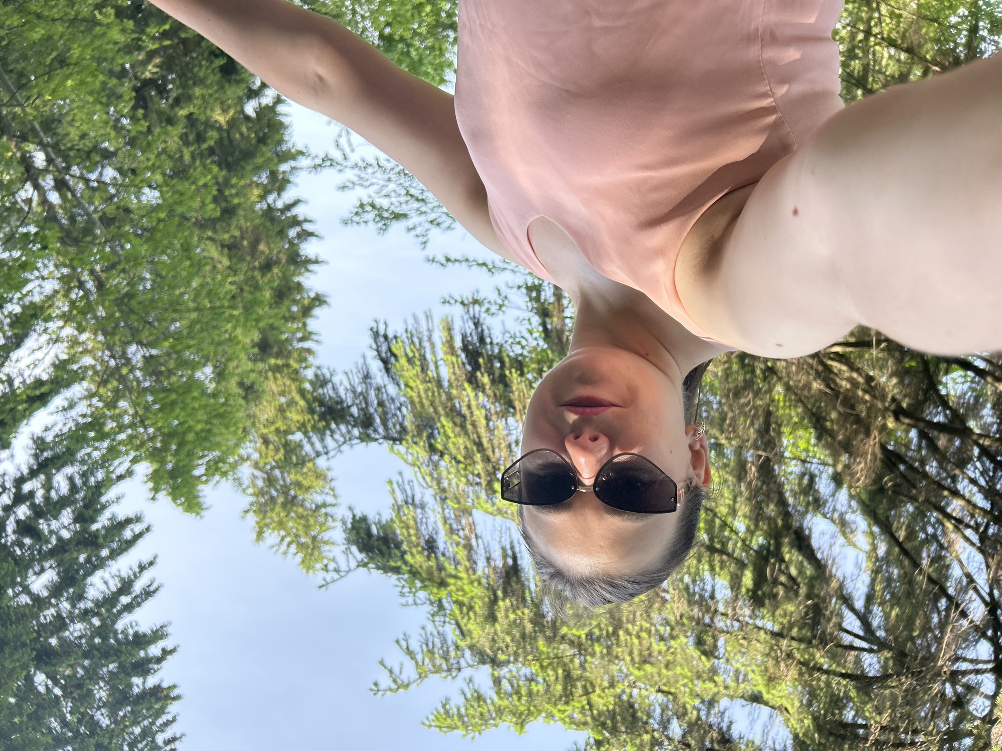 A selfie-style photograph shows a woman outdoors in Chaikovsky, Russia. She is wearing sunglasses and a pink top, with trees and sky visible in the background. The photo is taken from a low angle, looking up at her.