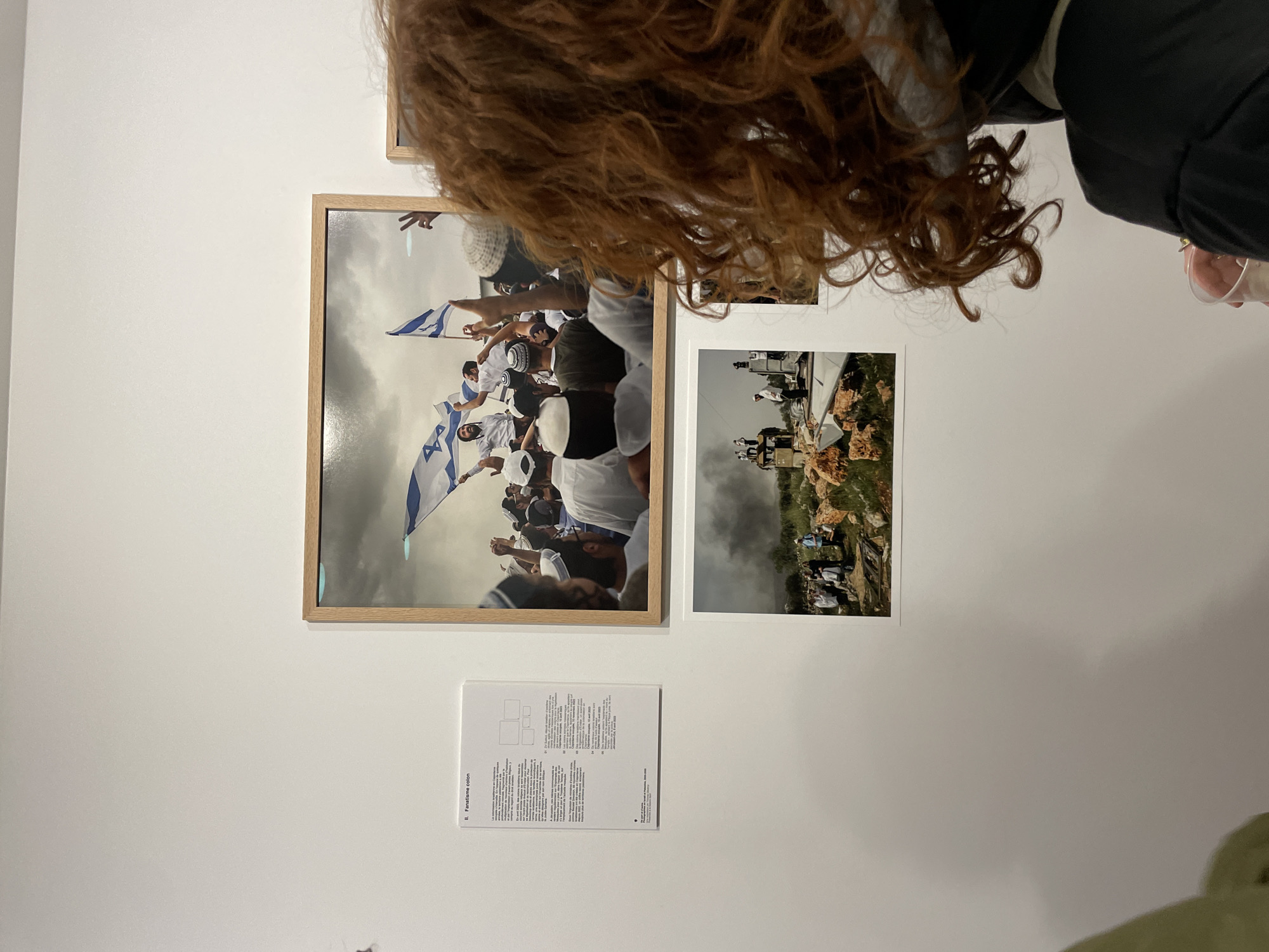 The image captures an indoor art exhibition at the Bourse de Commerce – Pinault Collection in Paris, France. A person with voluminous, curly reddish-brown hair, viewed from behind, stands observing a white gallery wall.
The wall displays a large, horizontally oriented framed photograph and a smaller, unframed photograph below it. To the left of the framed photograph, a white rectangular text panel titled "II. Fanatisme colon" (Colonial Fanaticism) in French provides contextual details, referencing Israeli settlers and the West Bank with specific dates.
The primary framed photograph depicts a densely packed crowd of predominantly male individuals, many wearing white kippahs, in what appears to be an outdoor setting. Several Israeli flags, featuring a blue Star of David on a white background with blue stripes, are prominently displayed. One bearded man is elevated above the crowd, holding an Israeli flag, while others raise their hands in gestures of celebration or demonstration under an overcast sky.
The smaller unframed photograph shows a group of individuals standing amidst rocky, uneven terrain with scattered debris and what appears to be demolished structures. Green vegetation is visible, and a cloud of smoke or dust rises in the background.
The observer in the foreground wears a dark jacket and holds a clear plastic cup containing a light-colored substance.