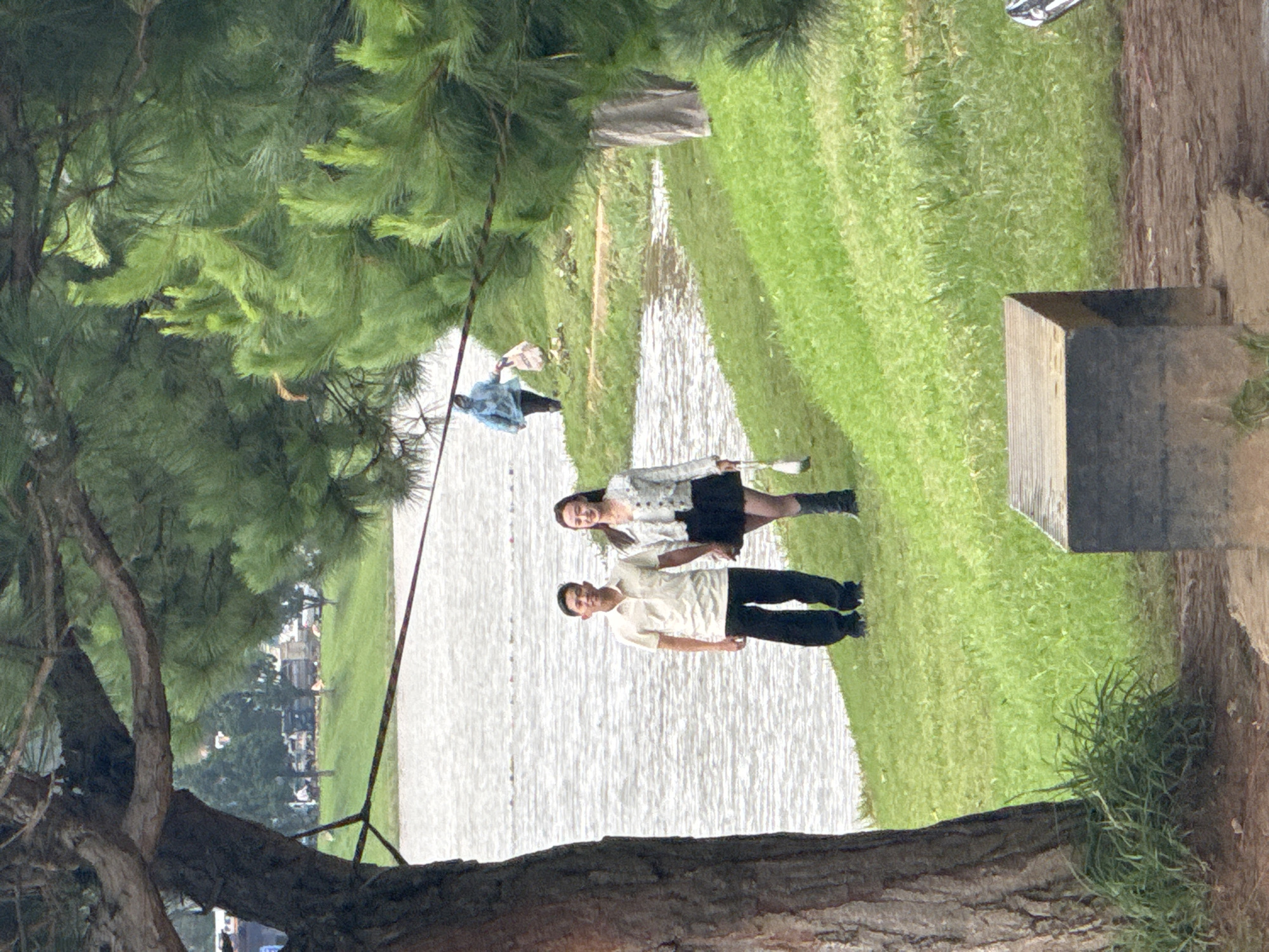 A daytime scene in Da Lat, Vietnam, features two individuals posing by a calm body of water, likely a lake or wide river. The image is captured from behind a large, gnarled tree trunk and branches that dominate the lower-left foreground, providing a natural frame. Dense green foliage, resembling pine needles, fills the upper-left of the frame.
In the mid-ground, a young man and woman stand on the grassy bank adjacent to the water, smiling towards the camera. The man, on the left, wears a light-colored short-sleeved collared shirt and dark trousers. The woman, on the right, is dressed in a white long-sleeved top with dark patterns, a black mini-skirt, and black boots, holding a thin, light-colored object in her right hand. Their relaxed postures and smiles suggest a pleasant occasion, possibly a photoshoot or a leisurely outing.
The expansive body of water extends through the center of the frame, reflecting the diffused light of an overcast or softly lit day. Green grassy banks line both sides of the water. Further back on the water, another person in a blue jacket is faintly visible. The right bank features a dark, cube-like wooden structure and some low-lying green foliage. In the distant background, across the water, faint structures and more trees can be discerned, adding depth to the tranquil landscape. There is no visible text in the image.