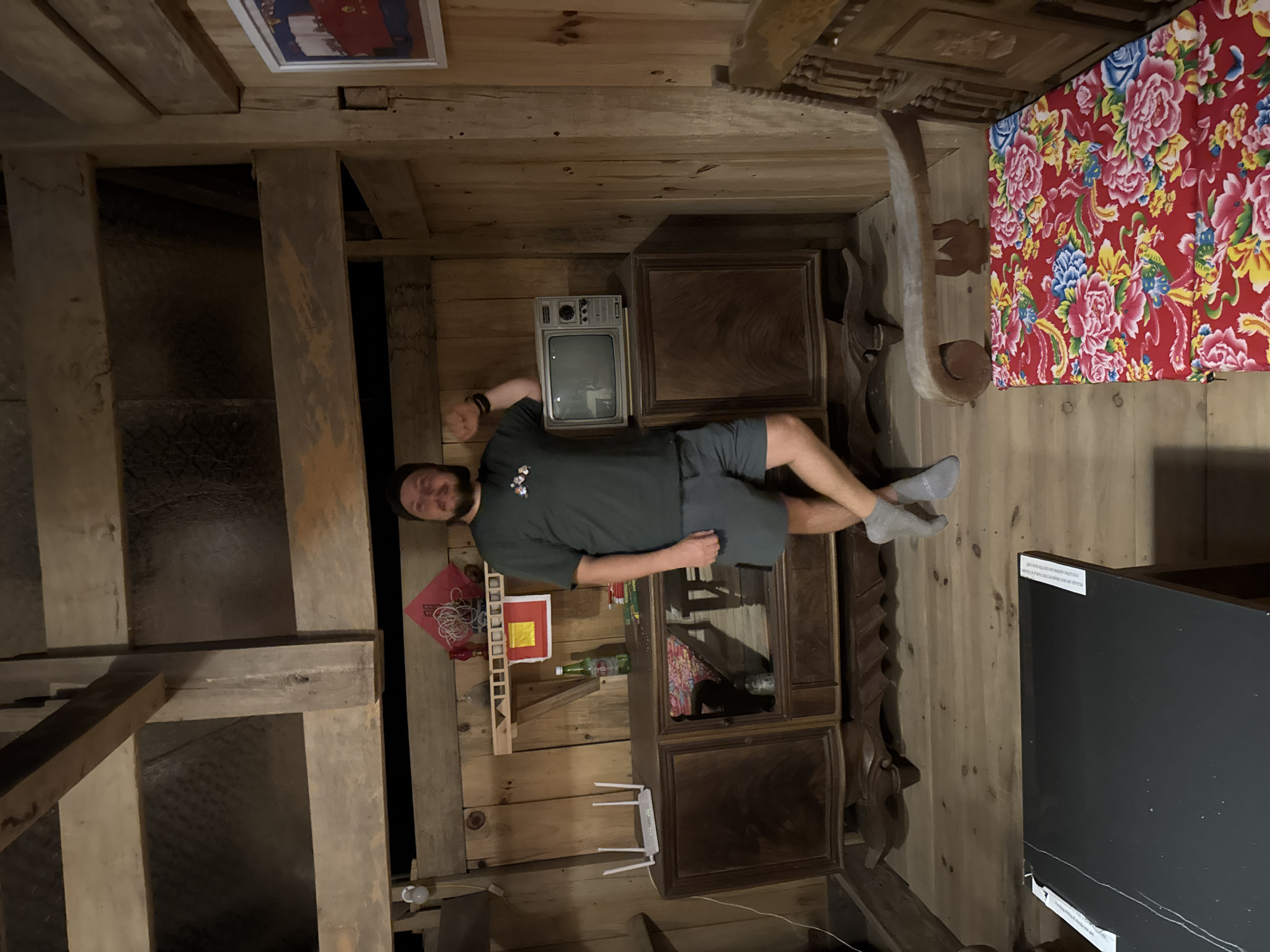 A horizontal shot reveals a man lying on an ornate, wooden cabinet in a room with wooden walls and ceiling. The location is the Hoàng Su Phì Commune in Vietnam.
The man is wearing a dark green t-shirt and grey shorts. He is lying on his side, facing the camera, with one arm outstretched.
The wooden cabinet is intricately carved and sits against a wooden wall. On top of the cabinet are a vintage television, a red flowered quilt, and a wooden sculpture. There are items on shelves in the cabinet including a router.
The room is lit with a warm, dim light, creating a rustic atmosphere. The ceiling is made of wooden beams, and the walls are also clad in wood. In the background, an opening in the wall is visible.