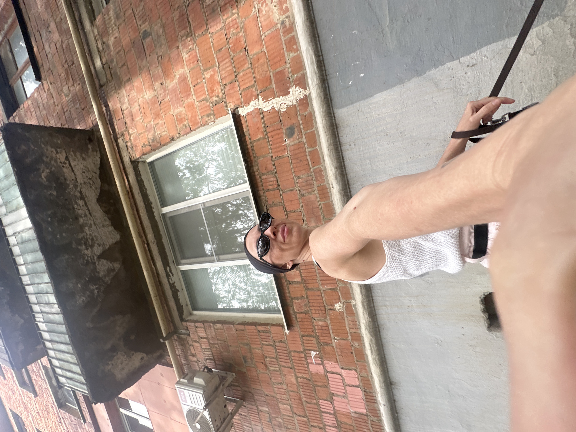 The image shows a low-angle, wide shot of a woman in a white tank top, pink shorts, and a black headscarf, wearing sunglasses. She is standing in front of a brick building with a balcony above a window. She is holding a dark leash in her right hand. The setting appears to be an urban environment. There is an air conditioning unit mounted on the exterior wall to the left of the woman. The building's facade is constructed of red brick and features several windows. The ground level has a light gray concrete wall, with a darker gray shadow cast across it. The balcony above has a weathered, textured underside and a railing.